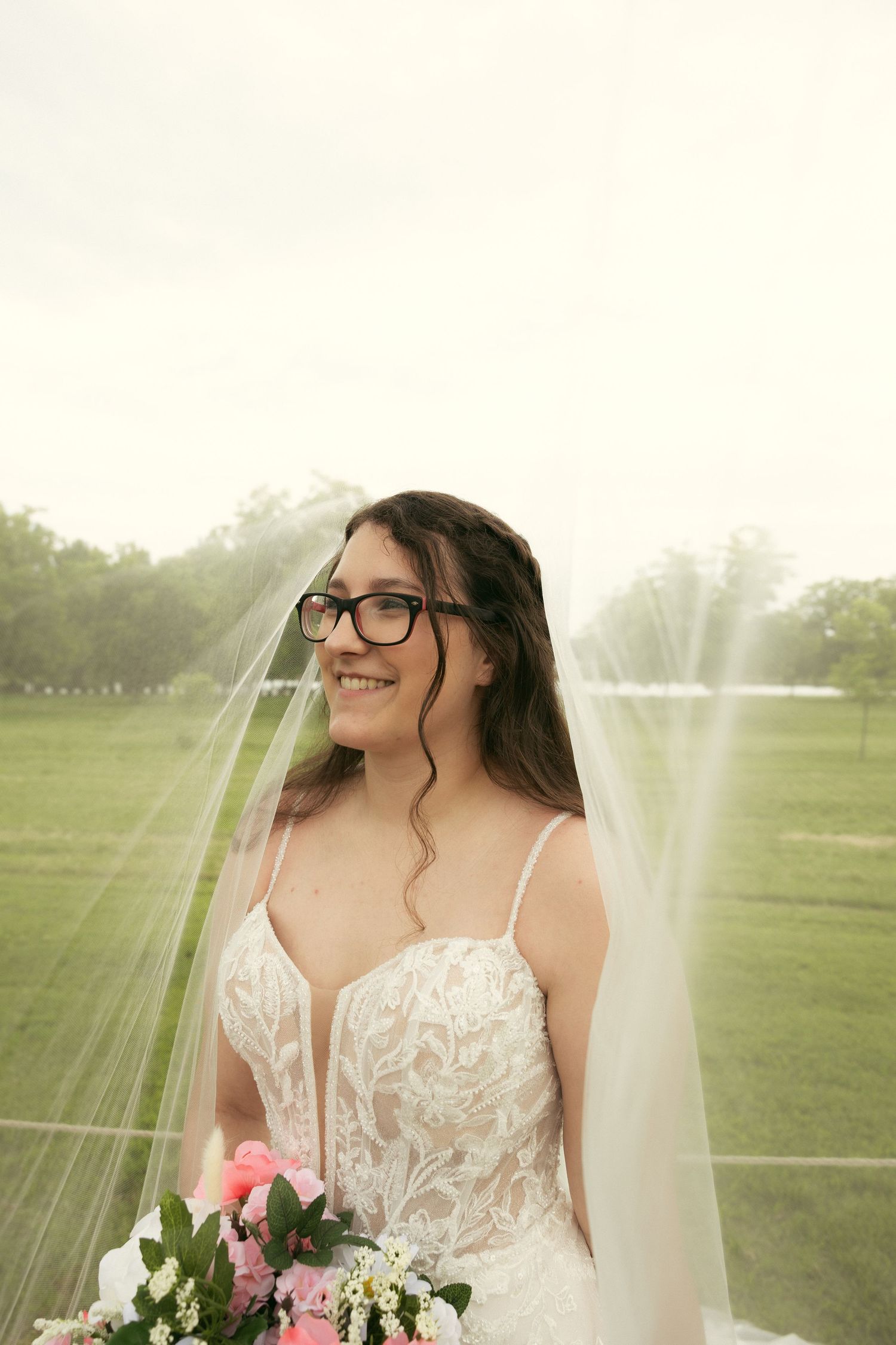 Smiling bride in glasses wearing a lace wedding gown and long sheer veil, holding a pink floral bouquet in a green grassy field.