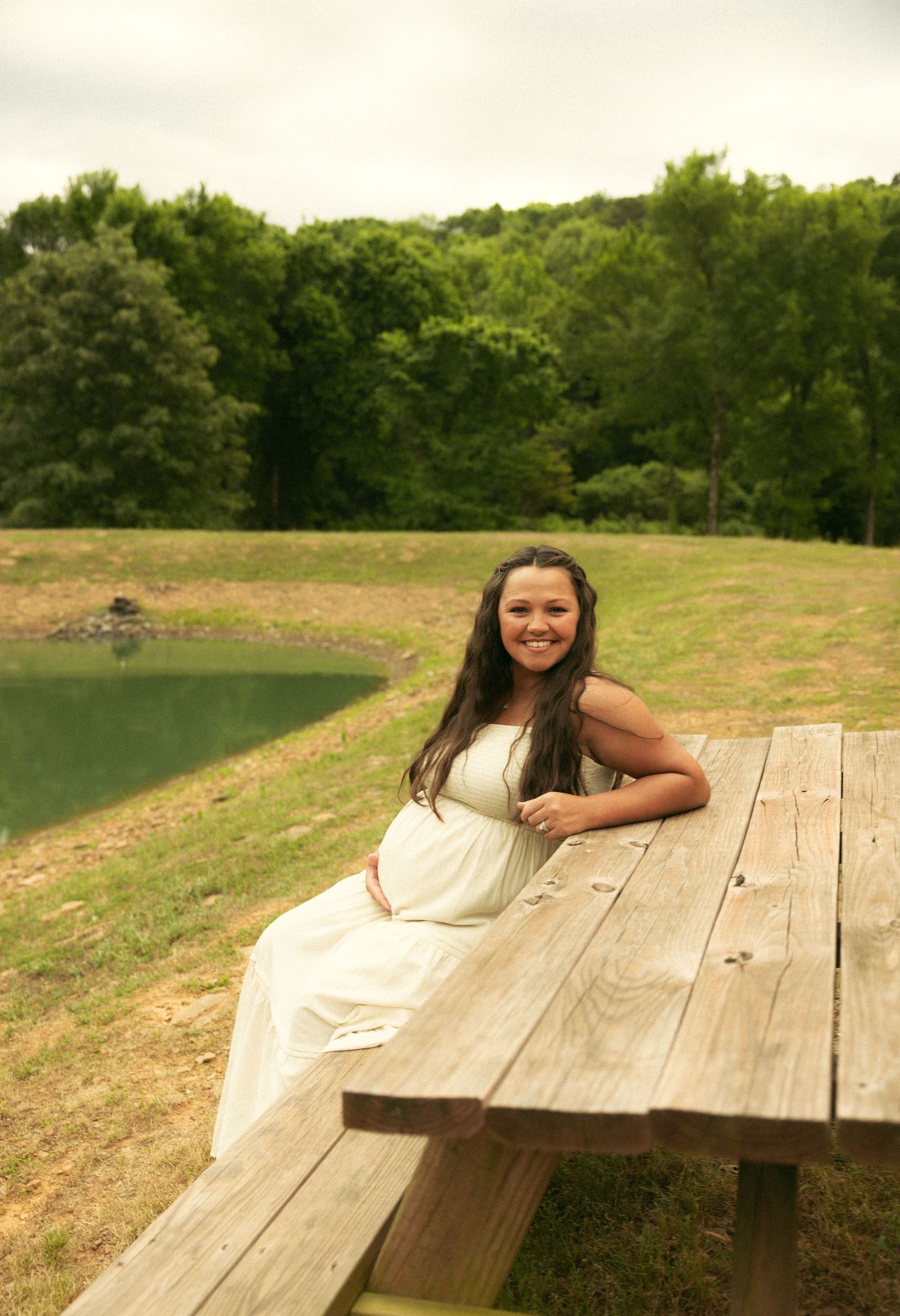 Smiling pregnant woman in white dress sitting at wooden picnic table by a pond surrounded by lush green trees.