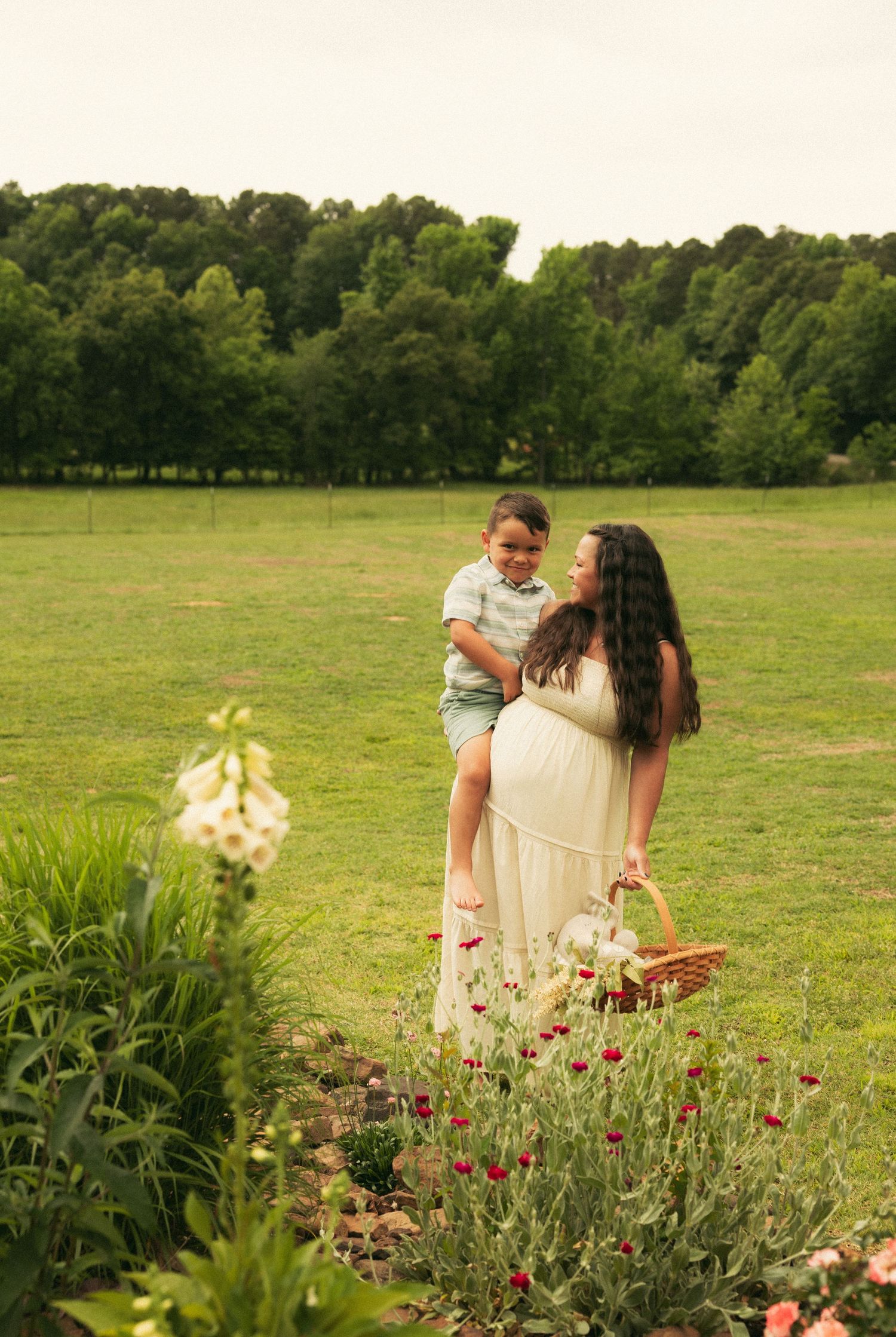 Pregnant woman in white dress holding basket lifting young boy in green field with wildflowers.