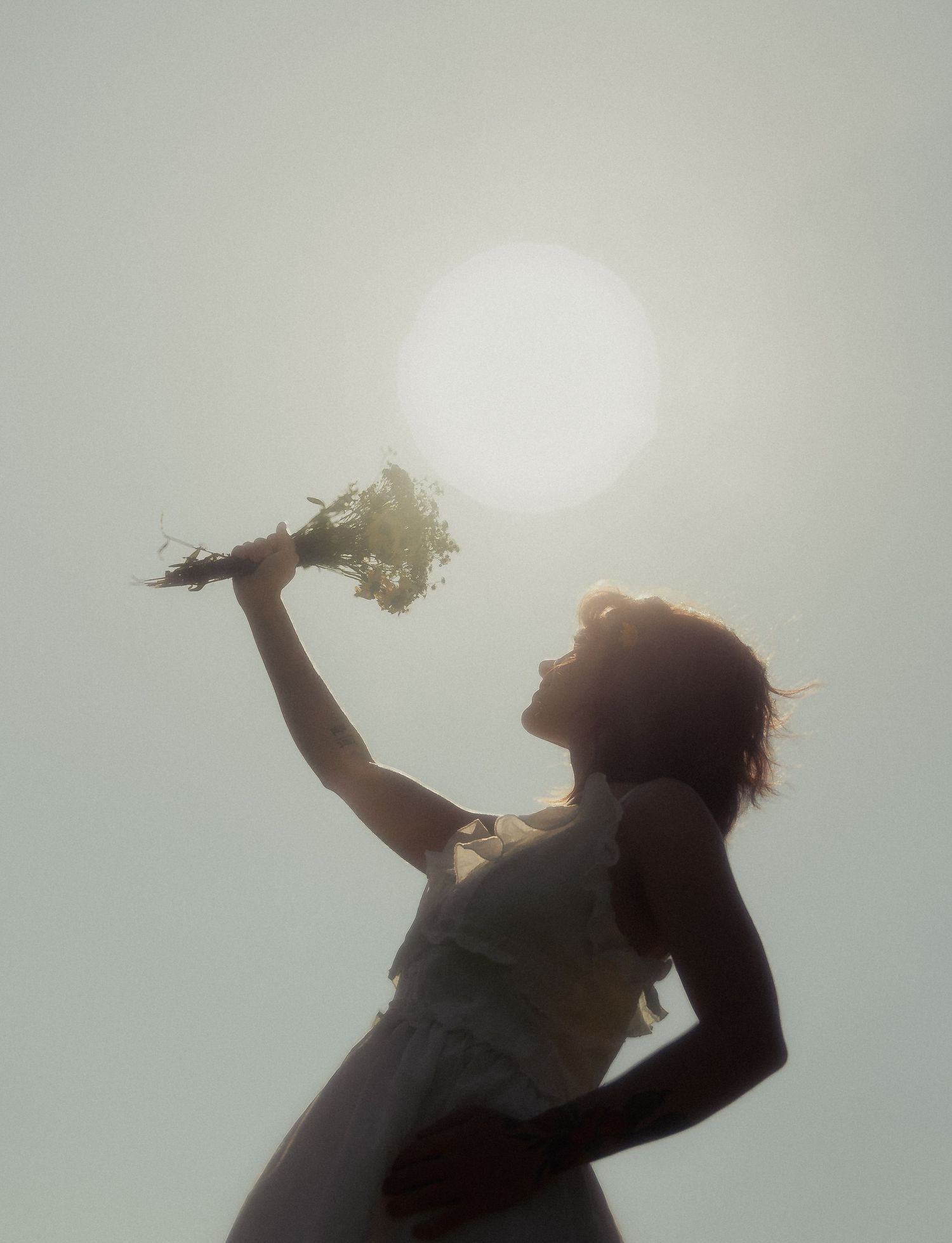 Silhouette of a woman in a white dress holding a wildflower bouquet up toward a bright glowing sun.