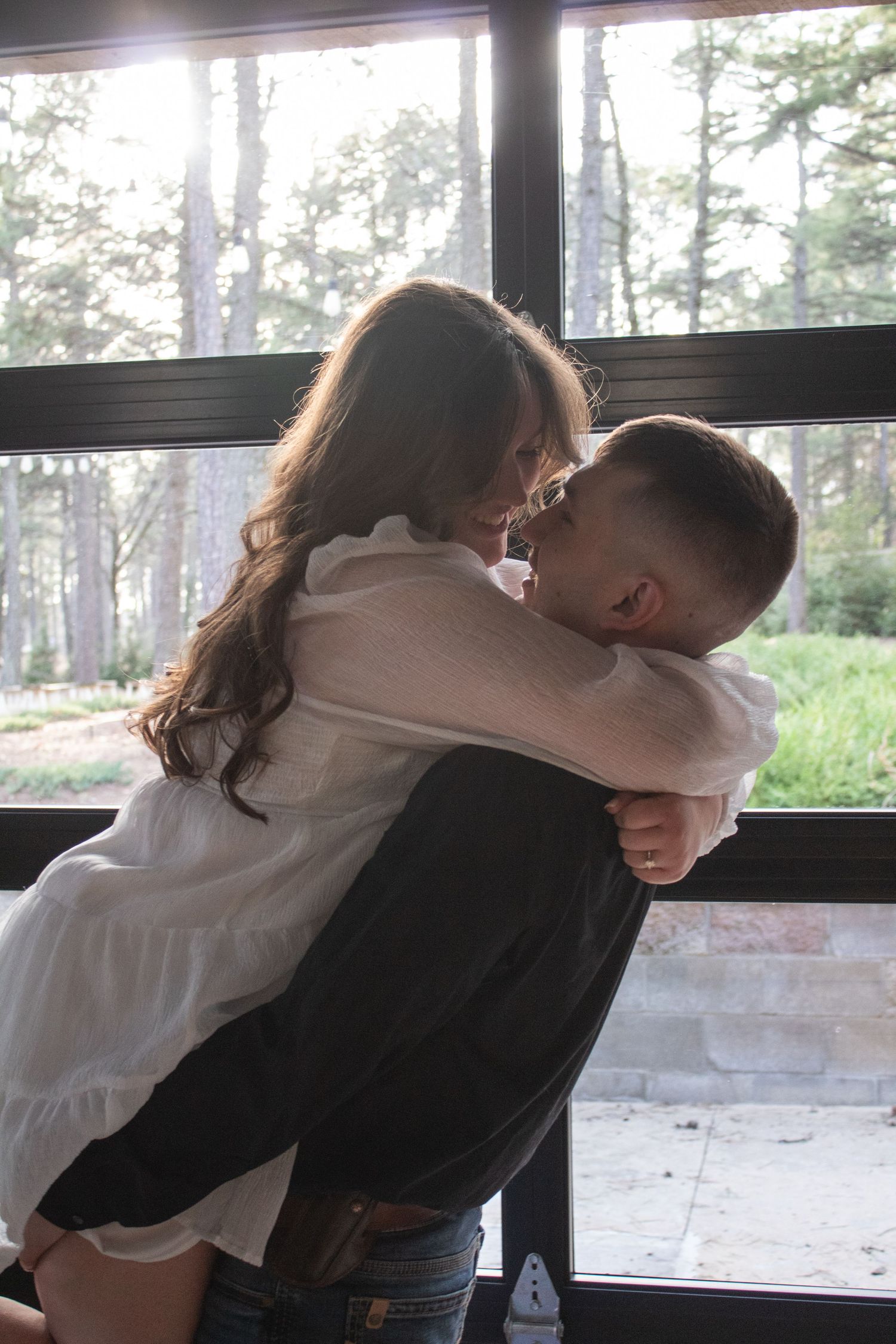 Couple sharing a tender kiss and embrace by a large sunlit window indoors.