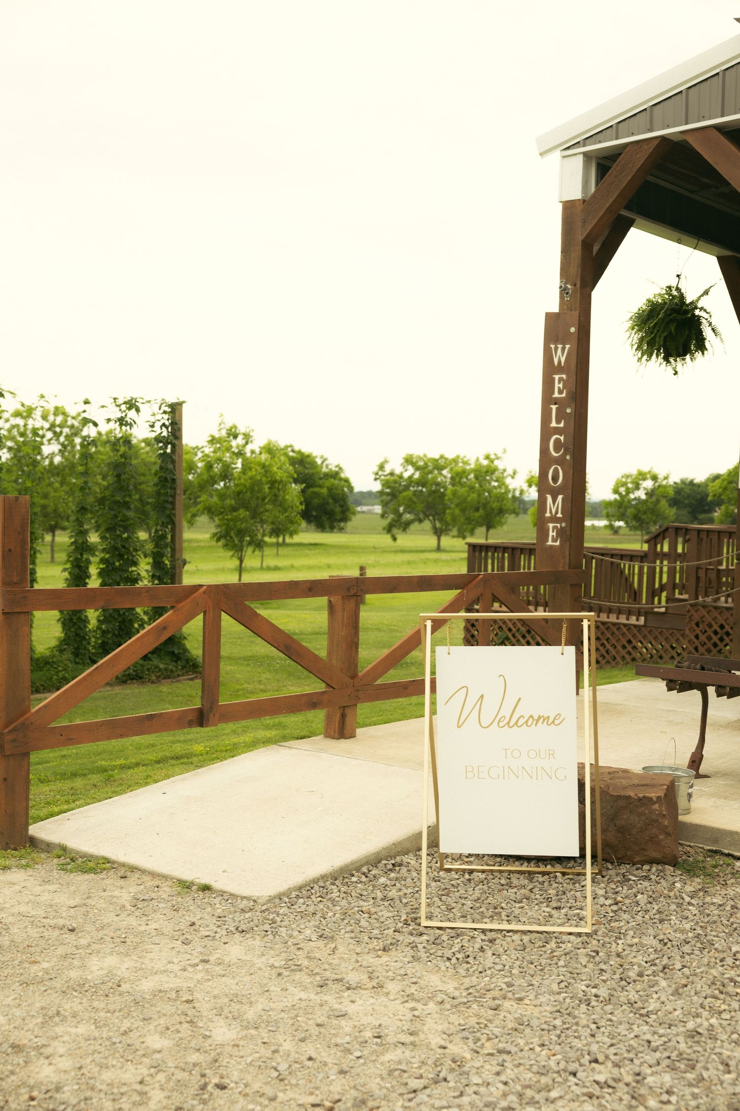 Rustic wooden welcome sign and white easel sign at a pavilion entrance with a wood fence and green farmland, a charming wedding venue.