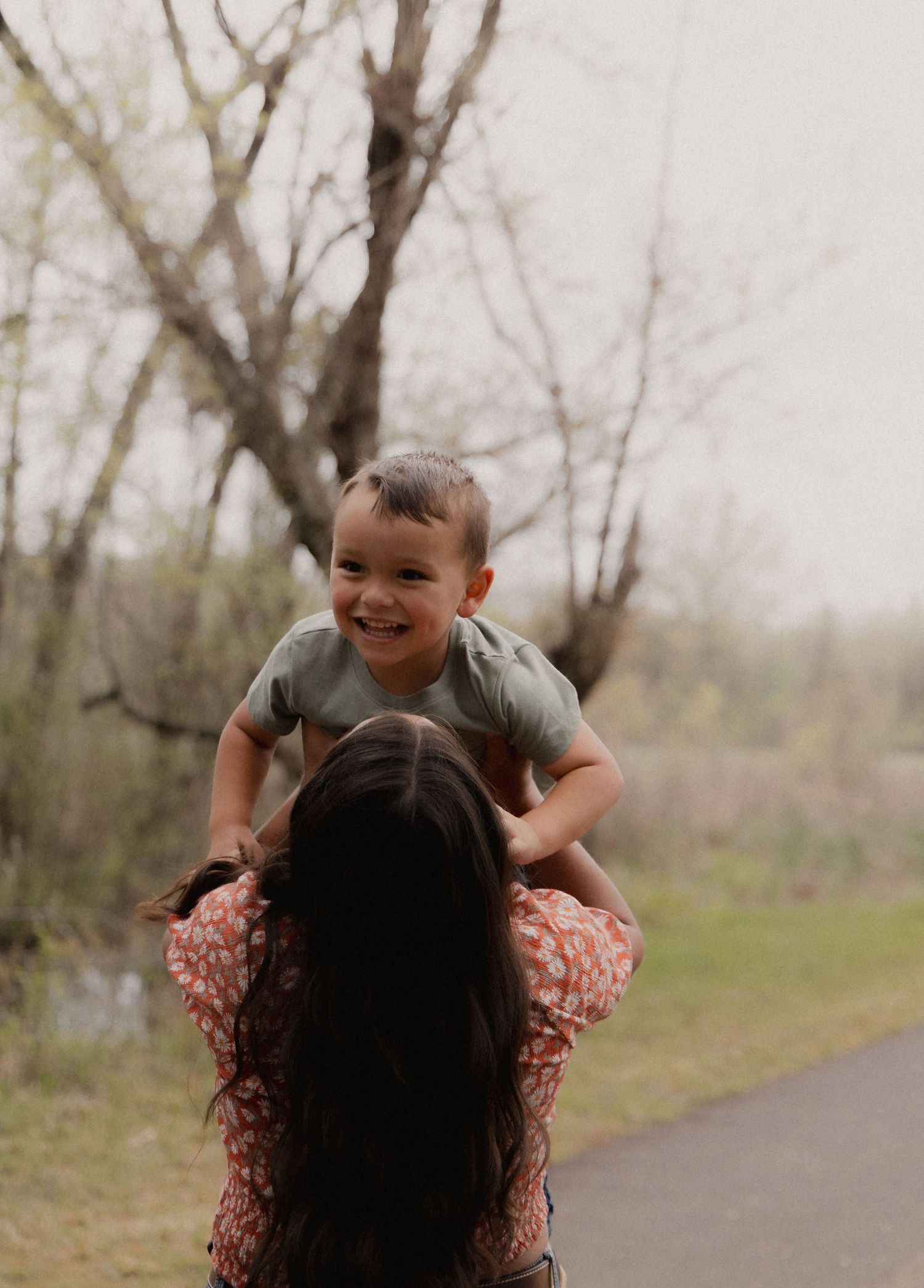 Laughing toddler boy being lifted overhead by his dark-haired mother on a nature path.