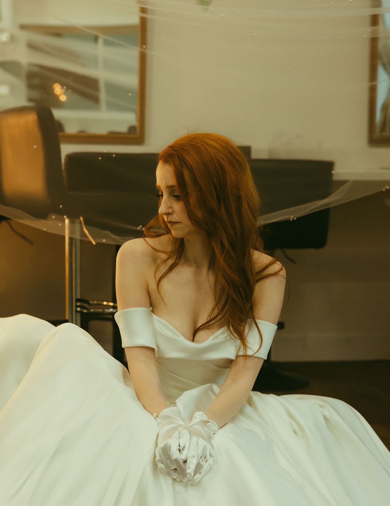 Elegant red-haired bride in off-shoulder white ball gown and gloves sitting thoughtfully in a bridal suite.