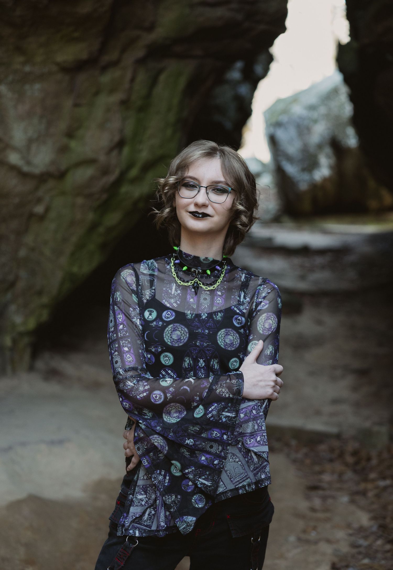 A young woman in a sheer cosmic-print top and glasses poses confidently inside a rocky cave formation.
