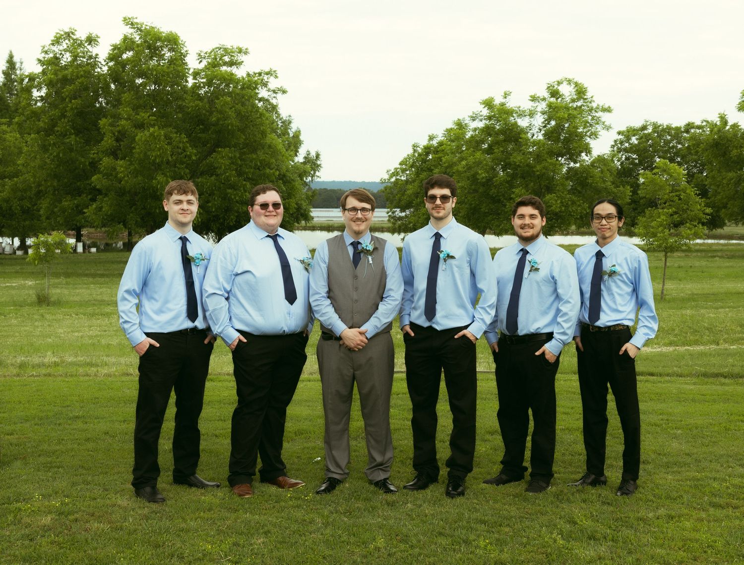 Six men in light blue dress shirts, dark ties and black pants pose side by side on a green lawn with trees in the background.