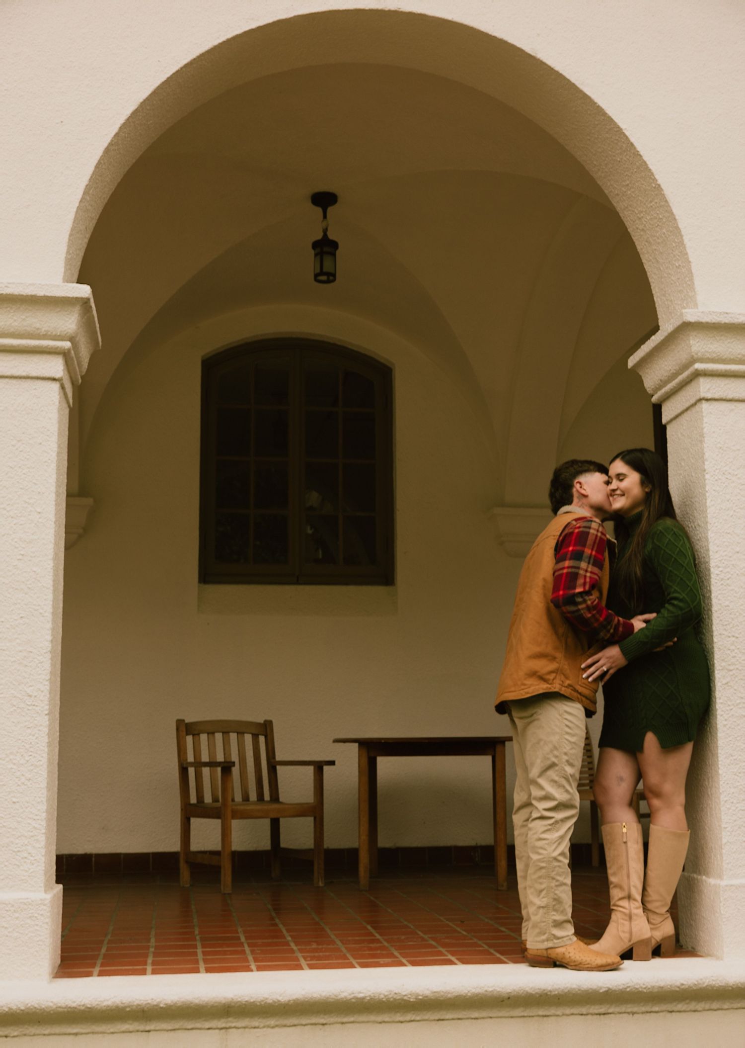 Romantic couple embracing under an arched portico with terracotta tile floor and wooden chair.