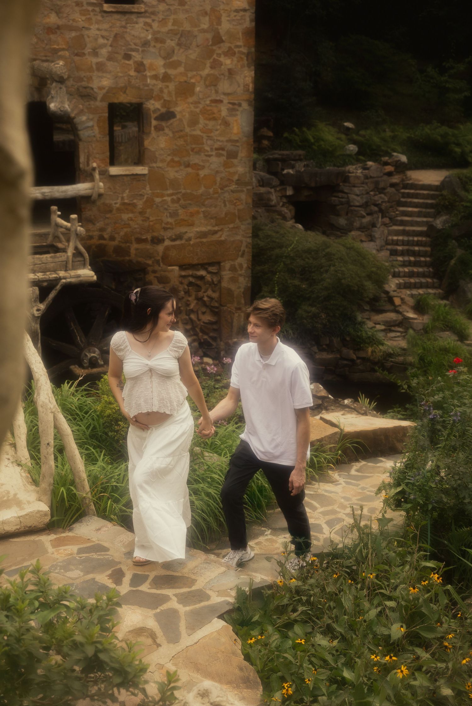 Expecting couple holding hands and walking along a stone path beside a rustic stone building.