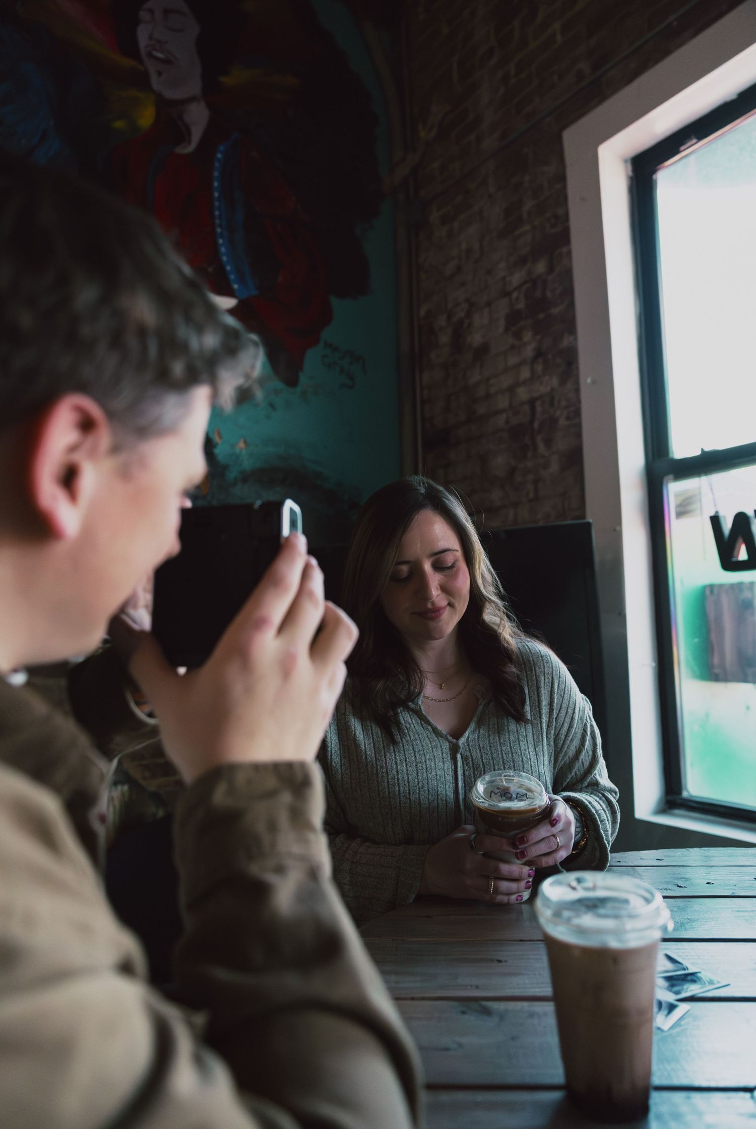 Couple sitting at rustic wooden table in a cafe, man photographing woman holding a dark iced coffee.