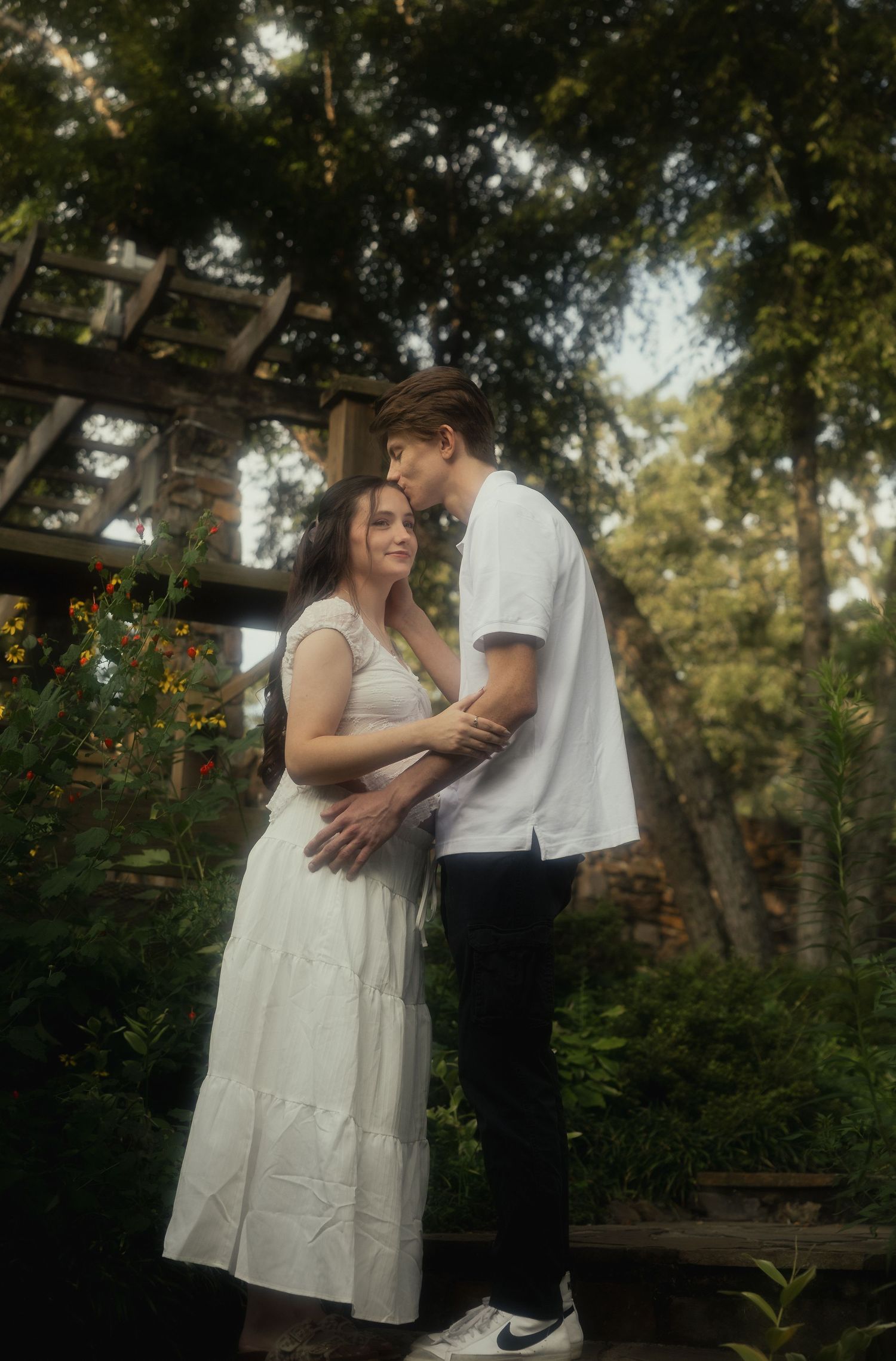 Young couple in white dress and white shirt embracing tenderly in a lush garden setting.