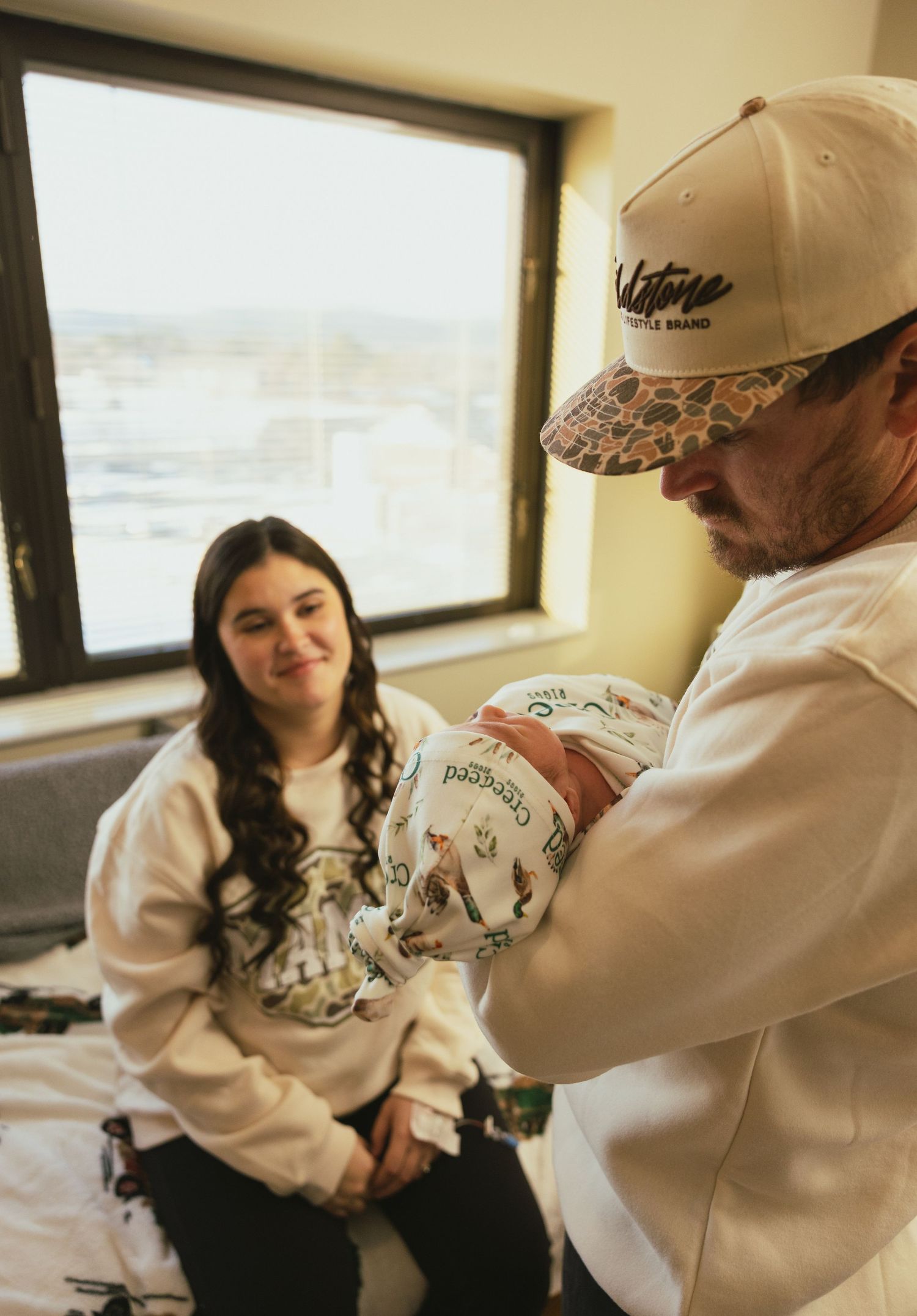A couple in a hospital room admires their swaddled newborn baby, the father wearing a tan cap and white hoodie.