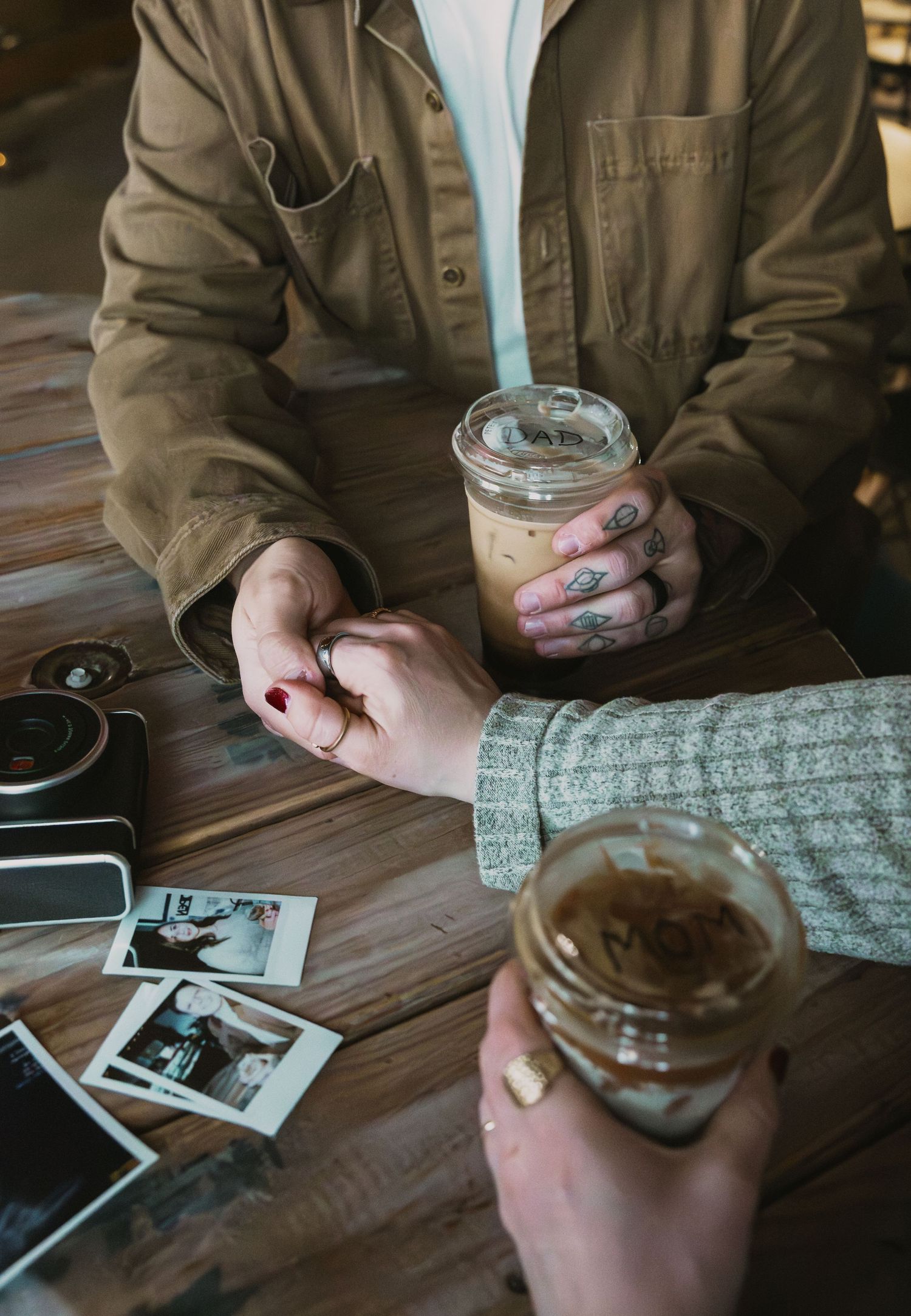 Couple holding hands at wooden table with iced coffees and Polaroid photos, man with tattooed fingers.