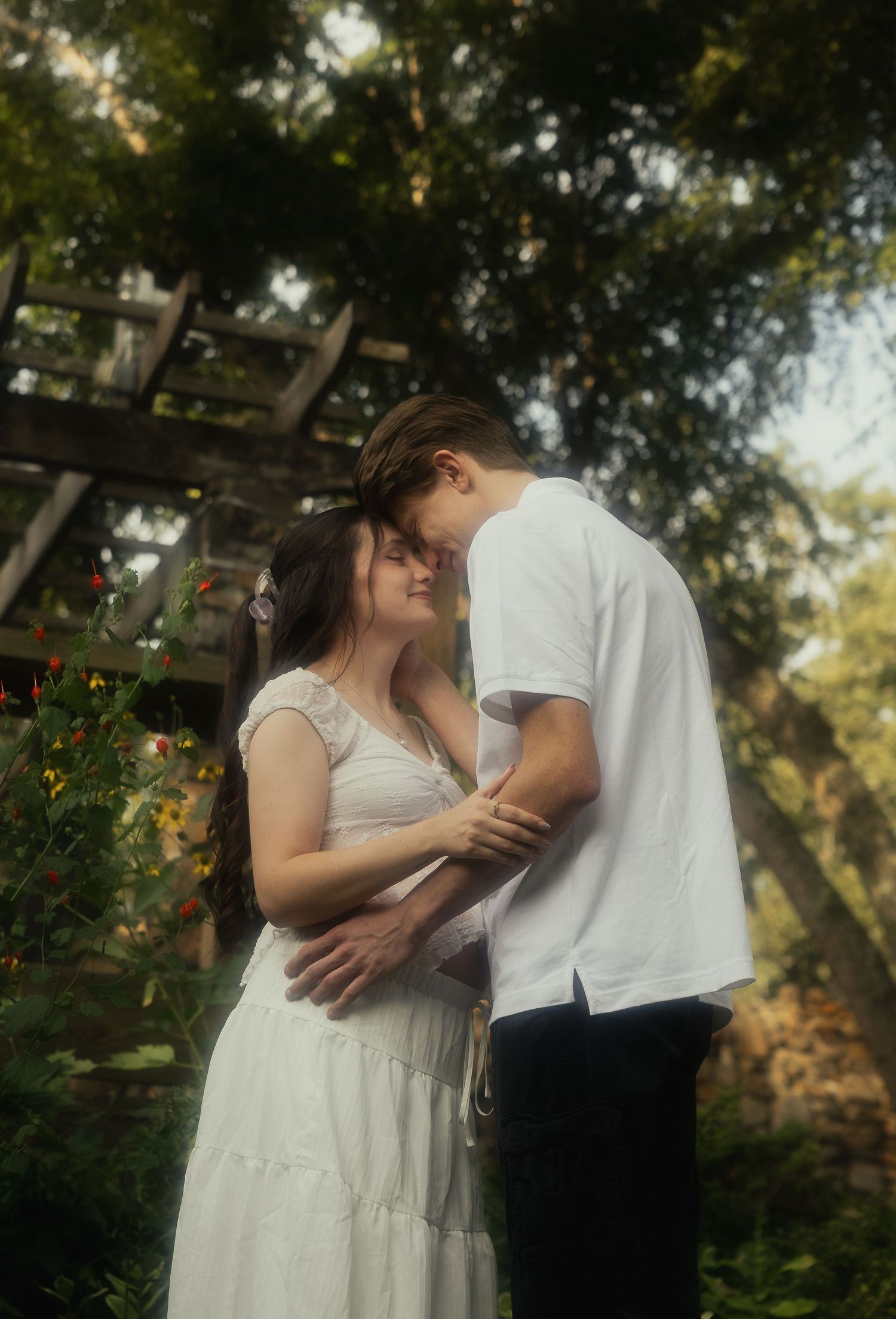 Couple in white outfits sharing a tender forehead-to-forehead moment under a garden pergola.