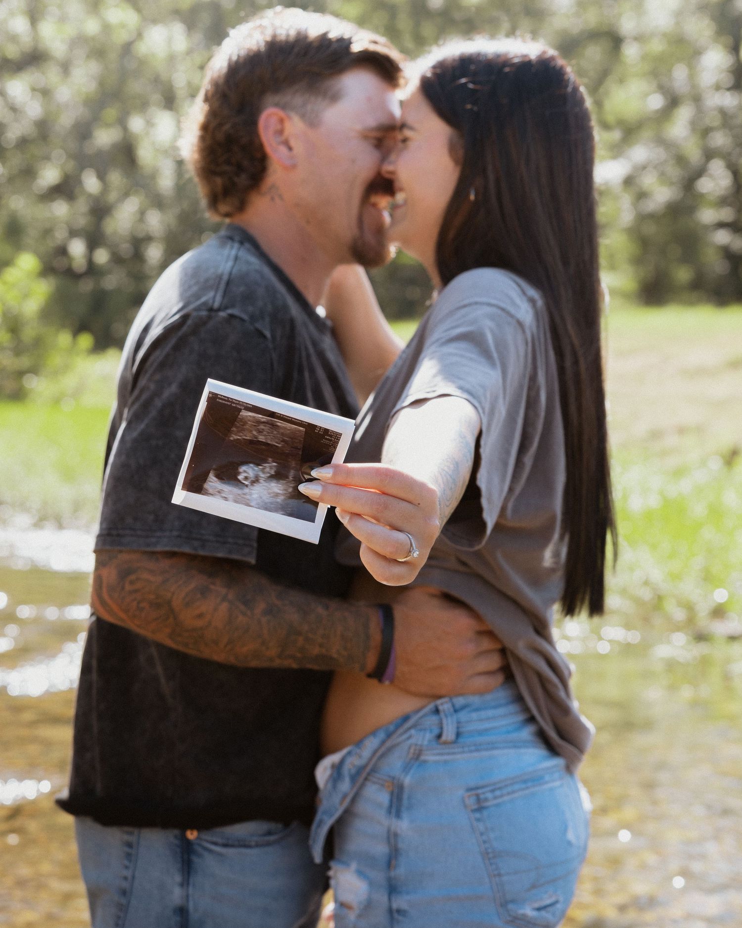 Couple kissing outdoors near water while holding ultrasound photo in sunny natural setting.