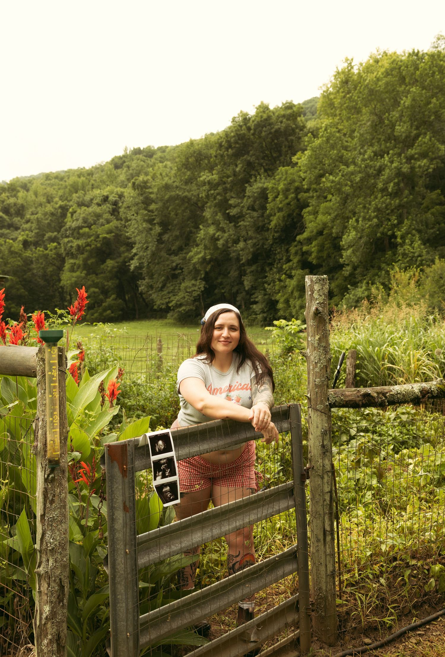 Pregnant woman leaning on rustic wooden fence in lush green farm setting, holding ultrasound photos, smiling outdoors.