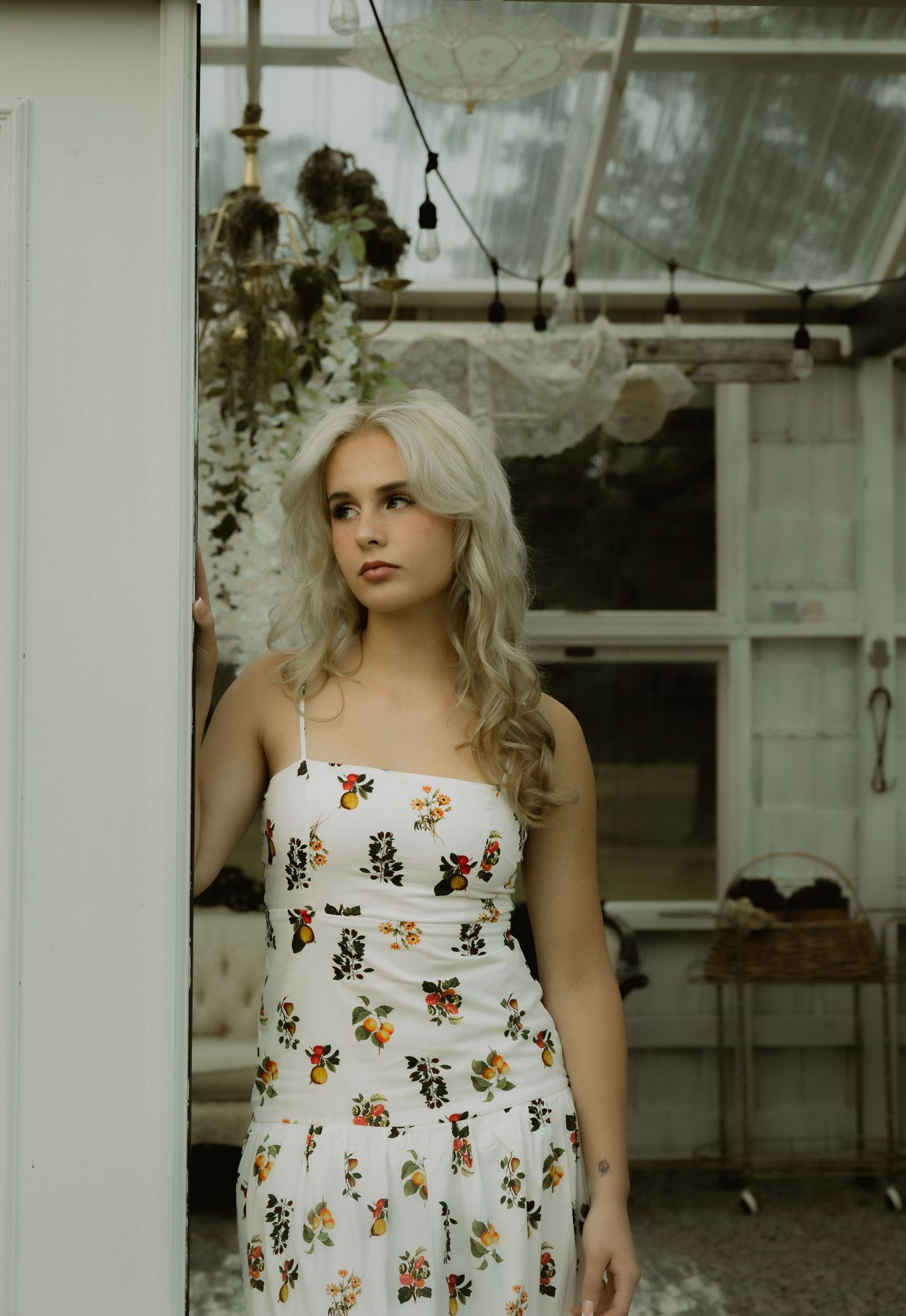 Blonde woman in a floral sundress stands in a doorway of a greenhouse with hanging dried flowers.