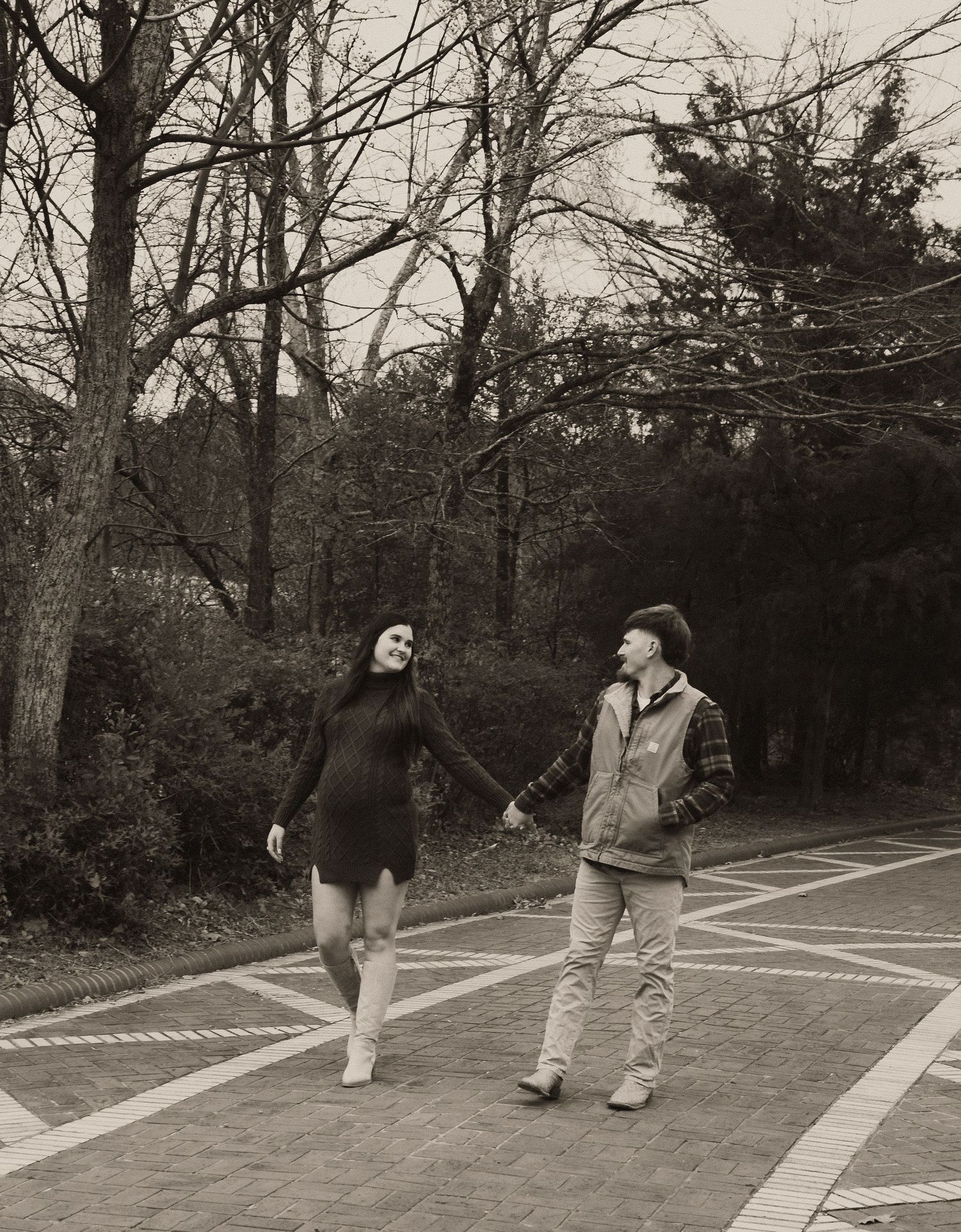 Couple holds hands walking on brick path in sepia-toned outdoor portrait among bare trees.