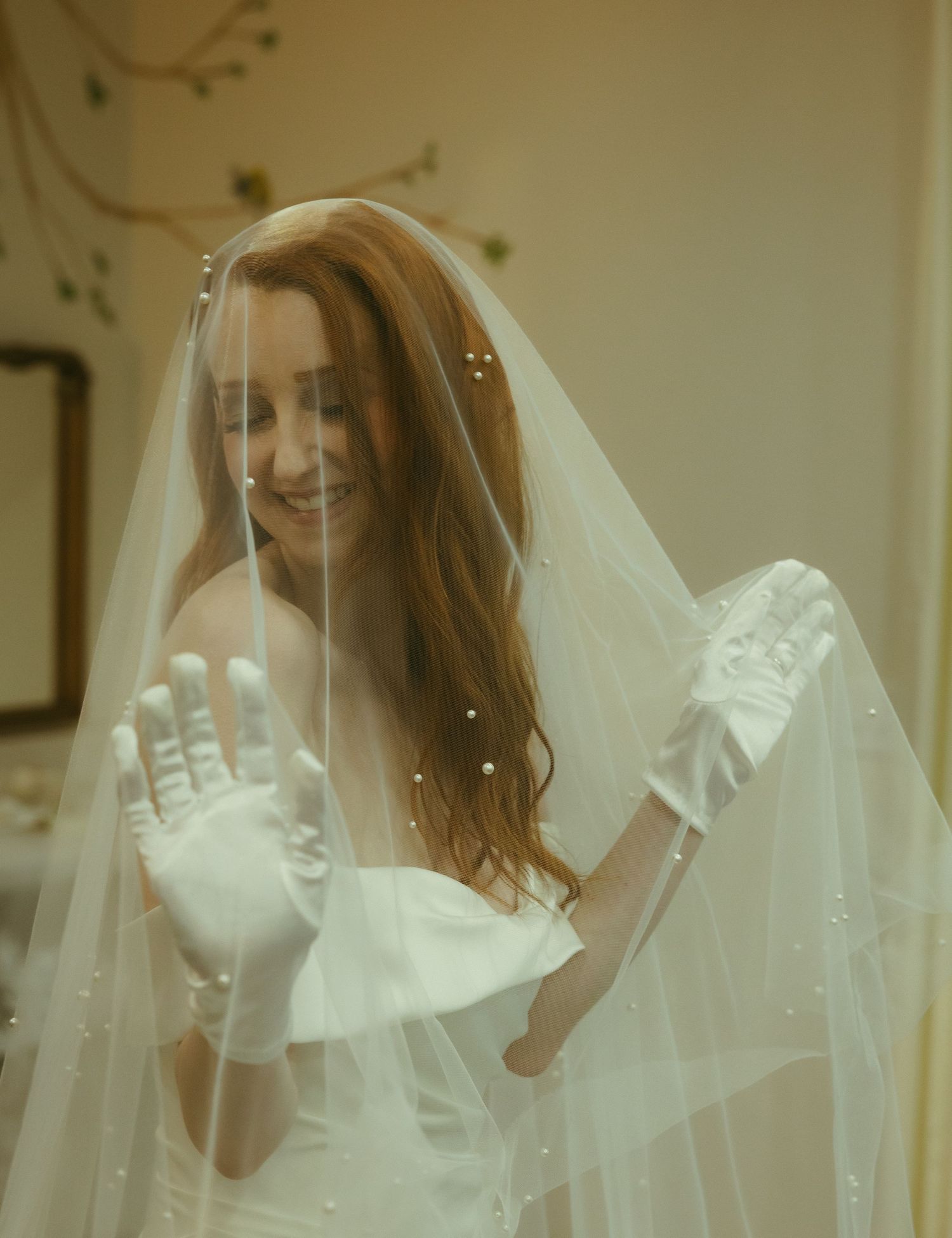 Smiling red-haired bride in white gown and veil holding up gloved hand during wedding preparations.
