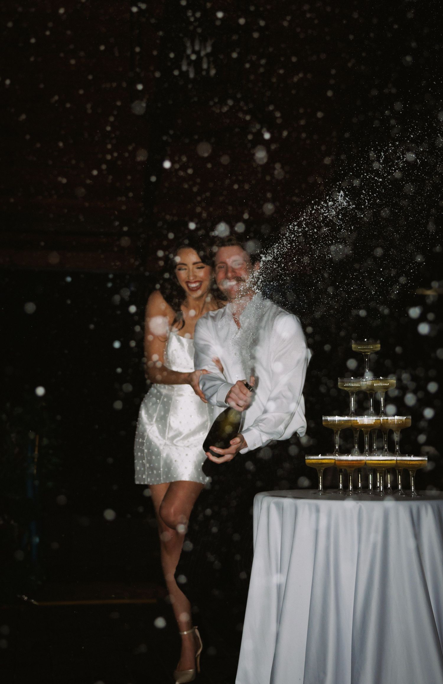 Couple celebrating with champagne tower spray at dark wedding reception venue.