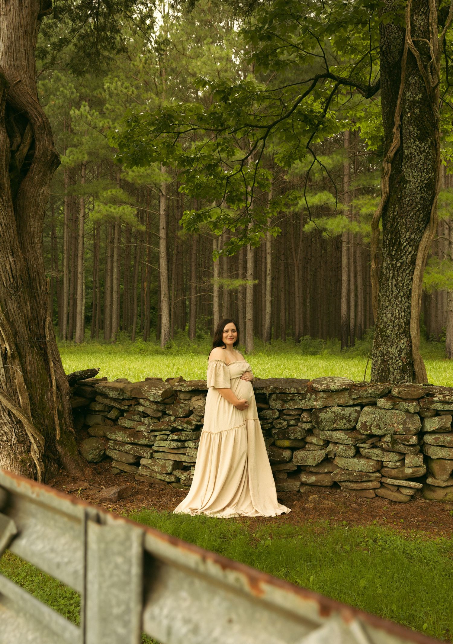 Pregnant woman in cream gown stands by stone wall in lush forest for an elegant outdoor maternity photo.