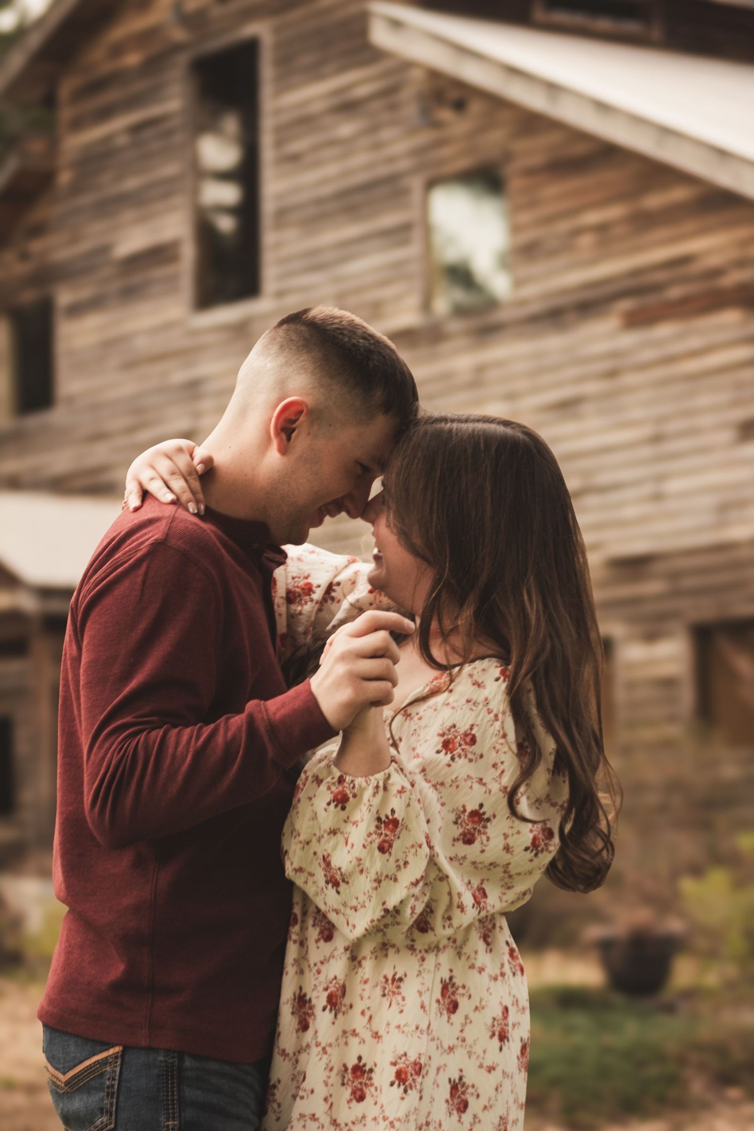 Romantic couple touching foreheads and holding hands in front of rustic barn.