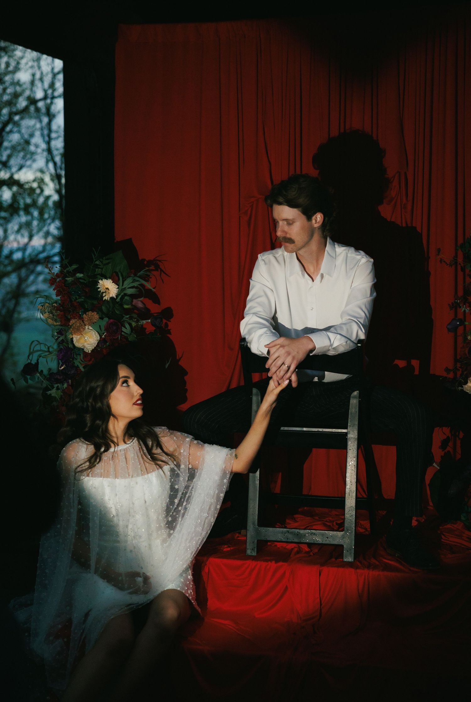 Couple in dramatic red-curtained room, woman in white lace dress seated while man in white shirt holds her hand.