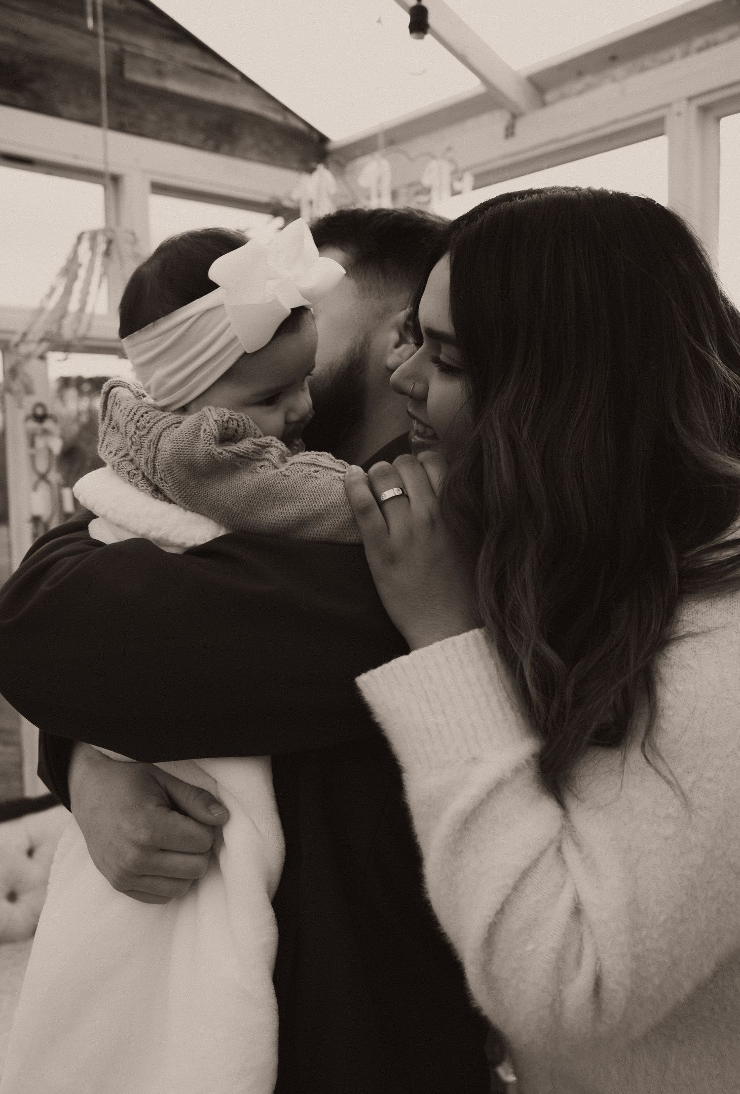 Black and white photo of parents kissing their baby girl with bow headband together indoors.
