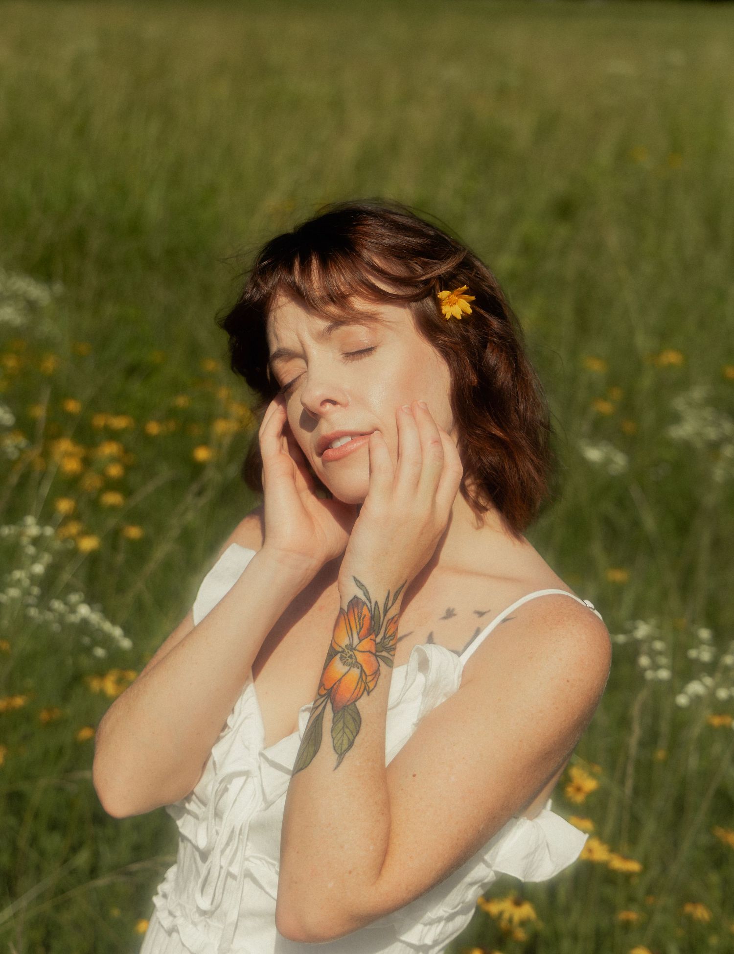 Young woman with flower in hair and floral tattoo poses peacefully in a sunny wildflower meadow.