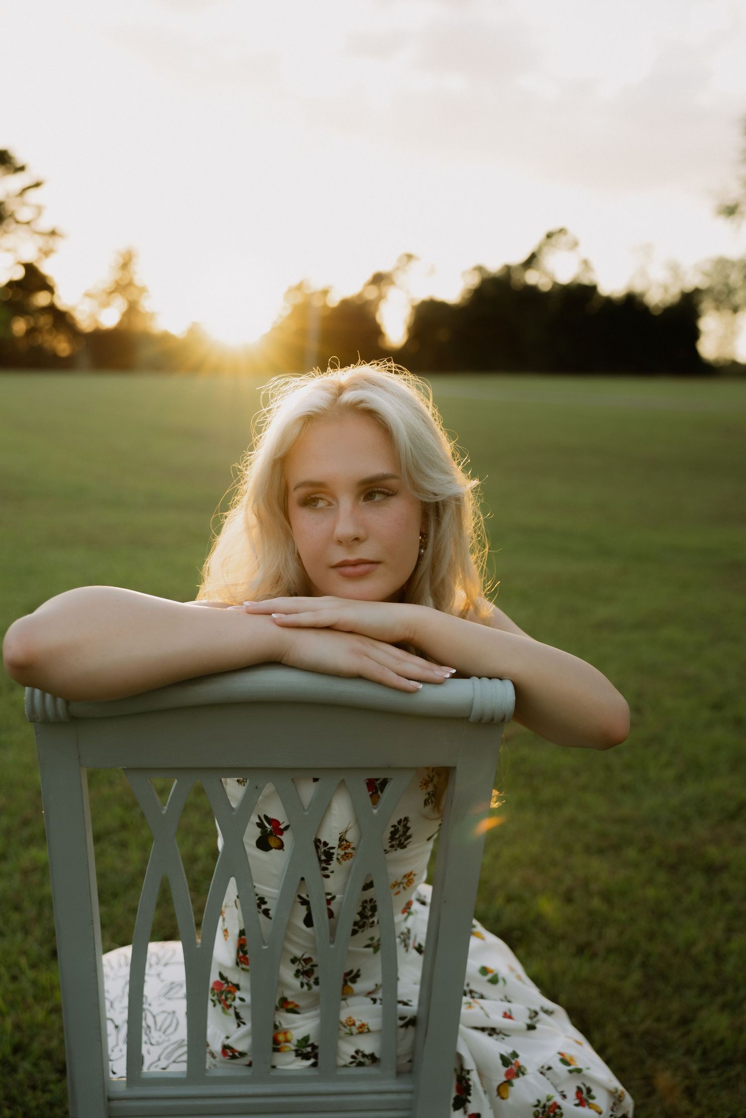 Young blonde woman resting chin on hands over chair back in a sunlit green field at sunset.