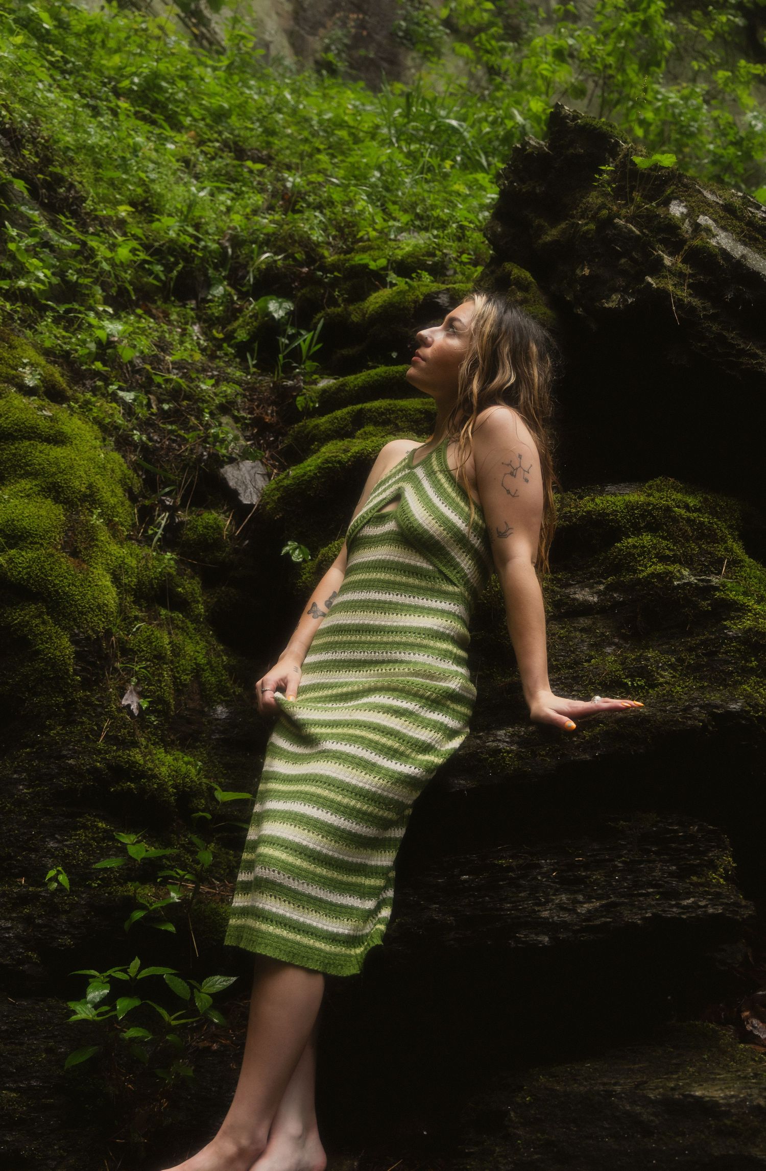 Woman in green striped knit dress posing against moss-covered rocks in a lush forest setting.