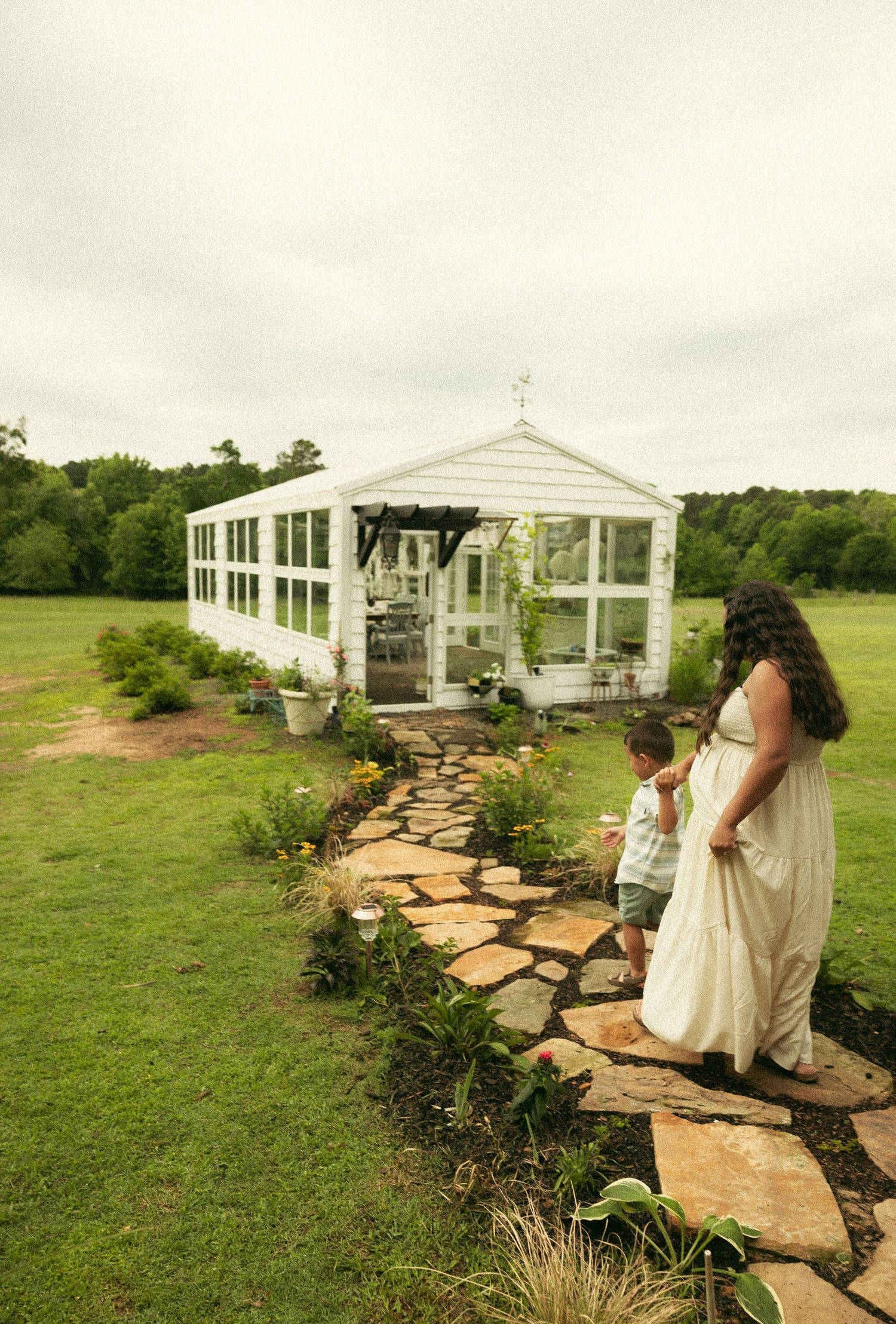 Pregnant woman in white dress walking with young child along stone path toward white greenhouse in green field.