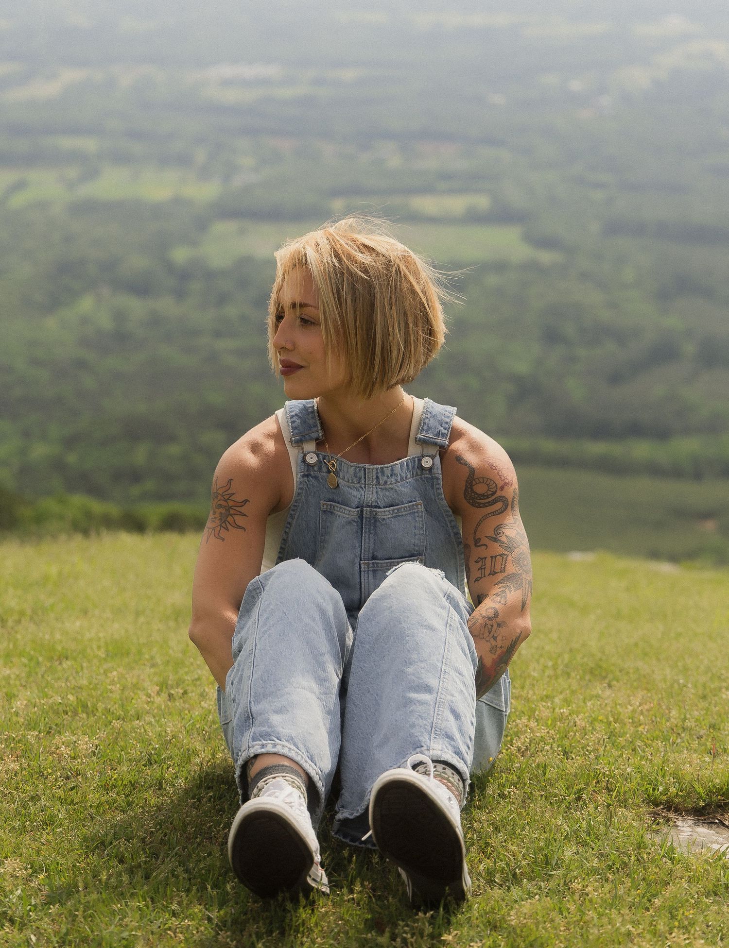 Tattooed blonde woman in denim overalls sitting on grassy hilltop with scenic view.