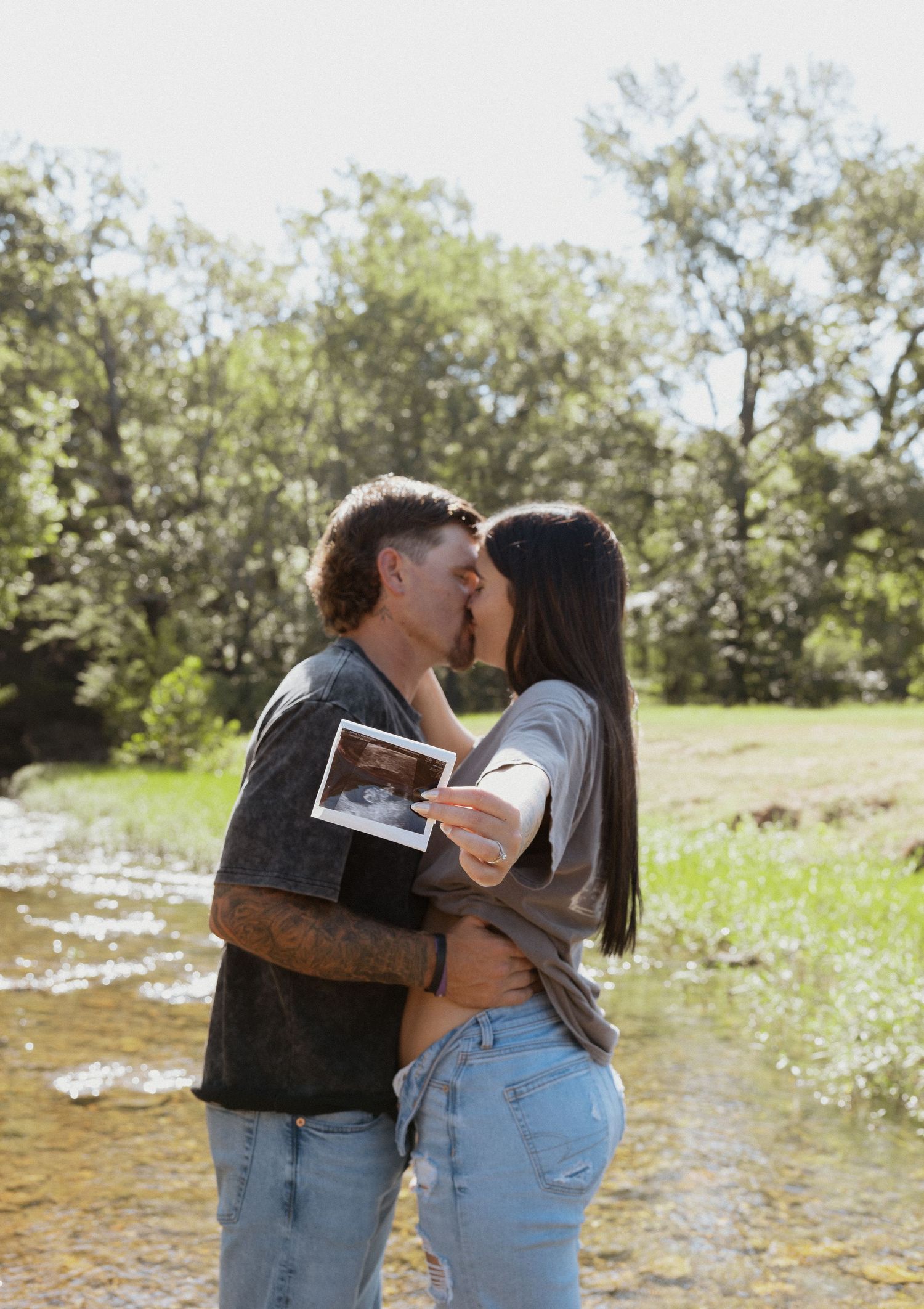 Couple kissing outdoors by a creek while holding an ultrasound photo in a sunny green setting.