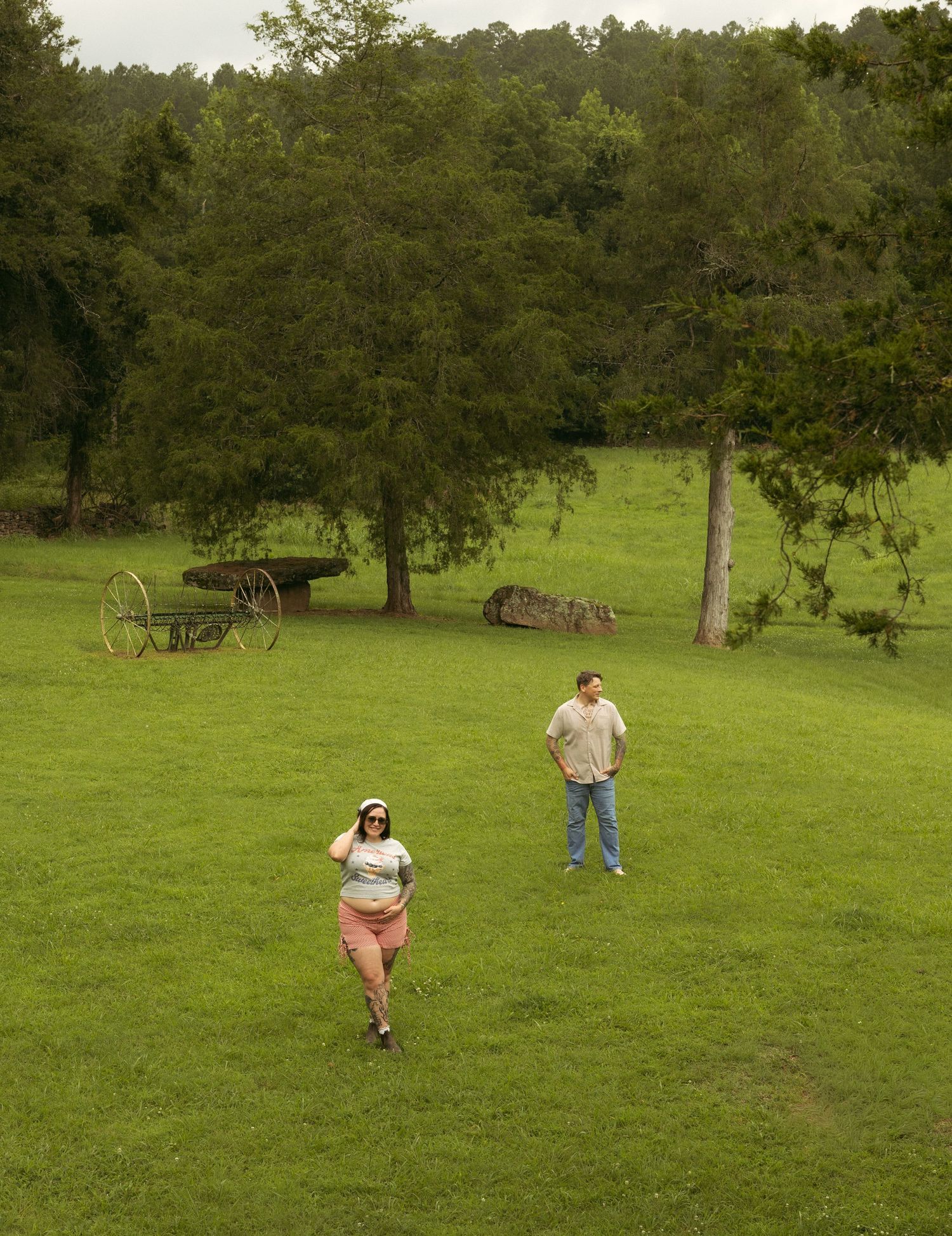Two people walking on a lush green farm field with vintage farm equipment and dense trees in the background.