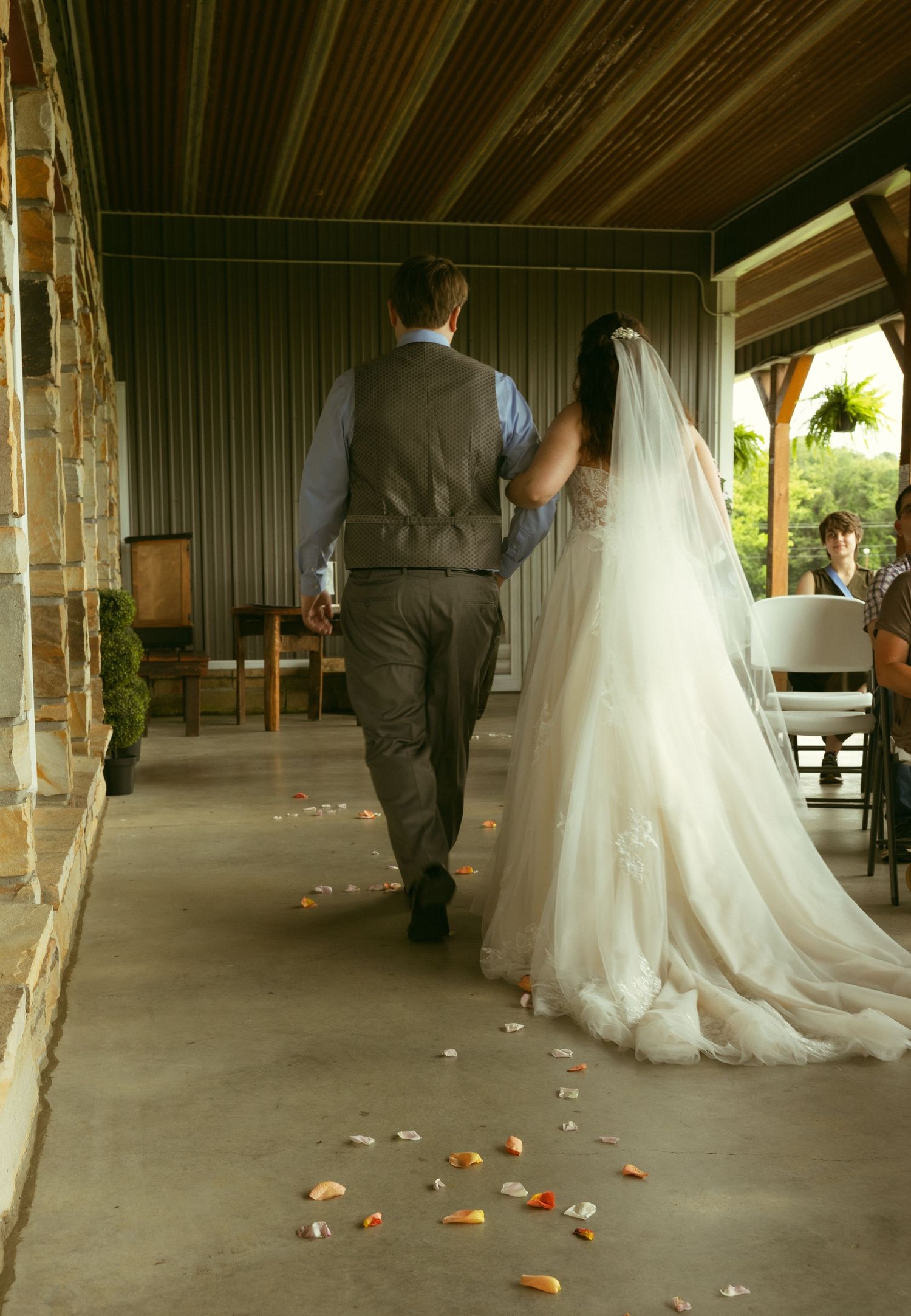 Bride in a long white lace gown and groom in a gray vest walk arm in arm down a rustic covered pavilion strewn with scattered flower petals.