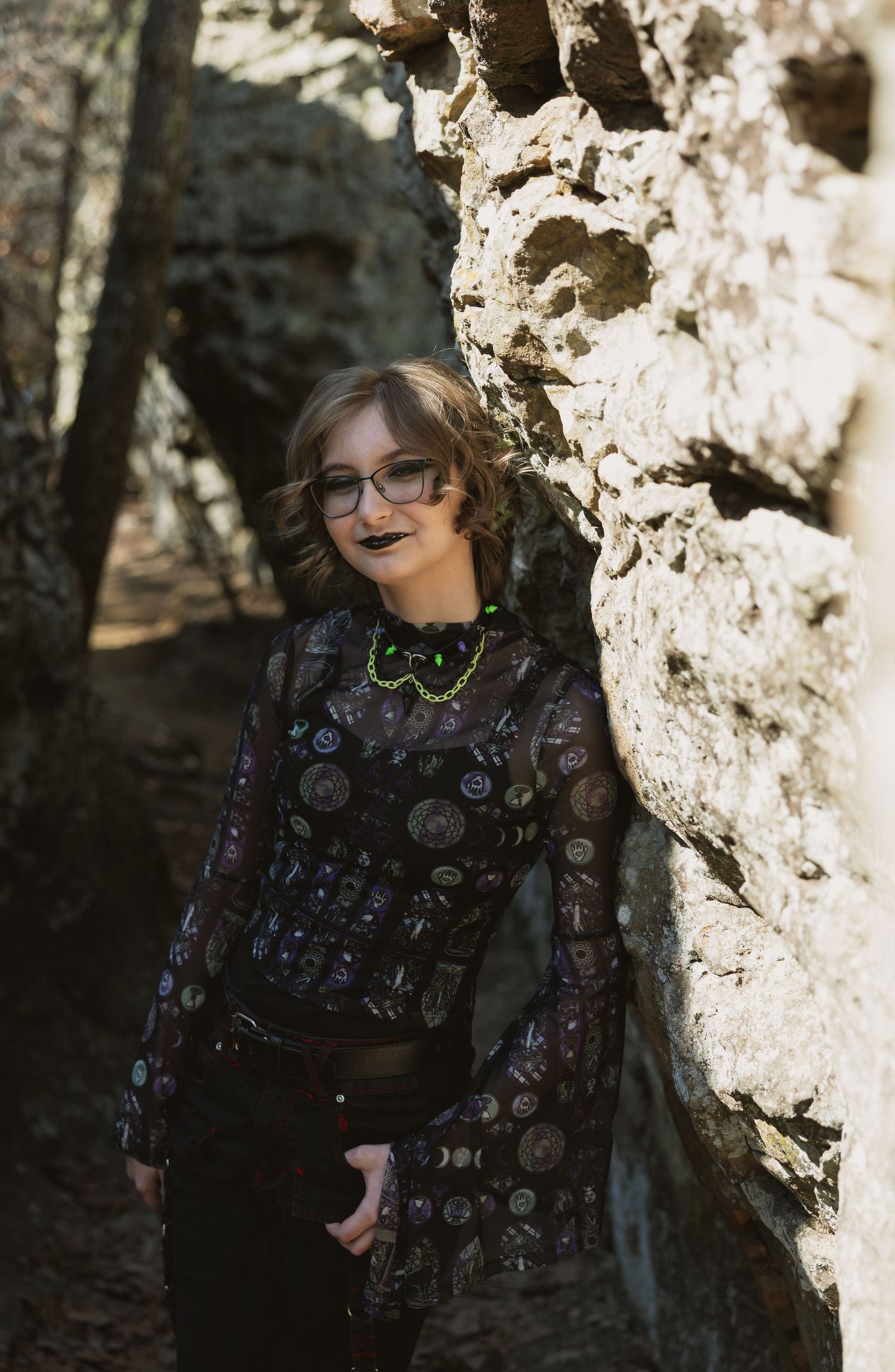 A young woman in a sheer patterned top and glasses leans against a sunlit rock wall in a natural setting.