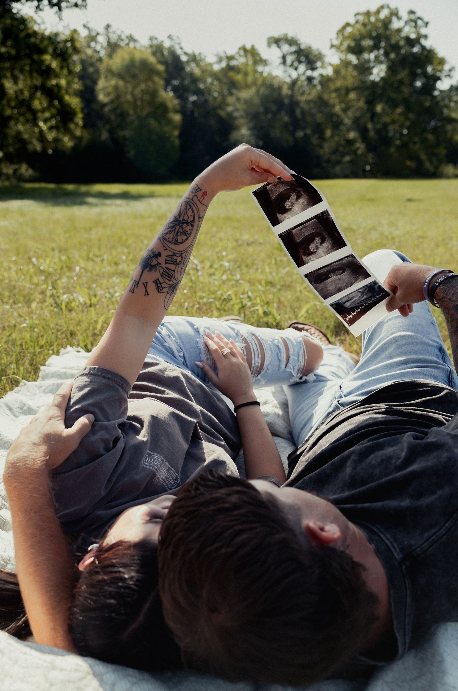 Couple lying on blanket in sunny park holding up ultrasound photos to announce pregnancy.