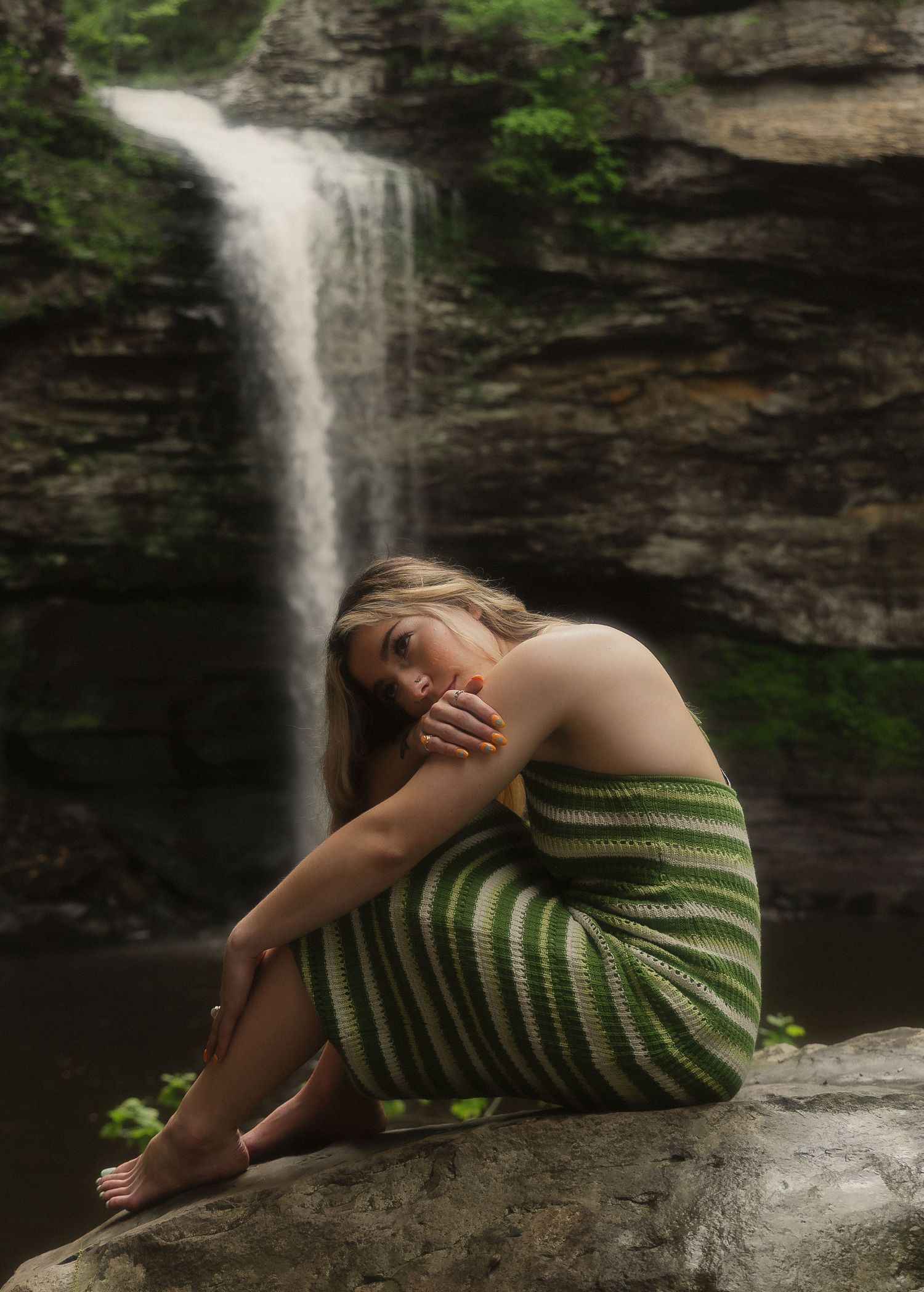 Young woman in green striped dress sitting barefoot on rocks in front of a cascading waterfall.