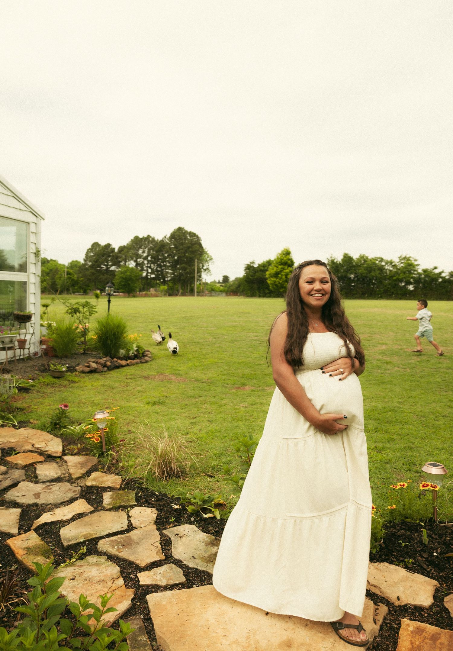 Happy pregnant woman in white dress cradling baby bump while standing on stone path in lush garden.