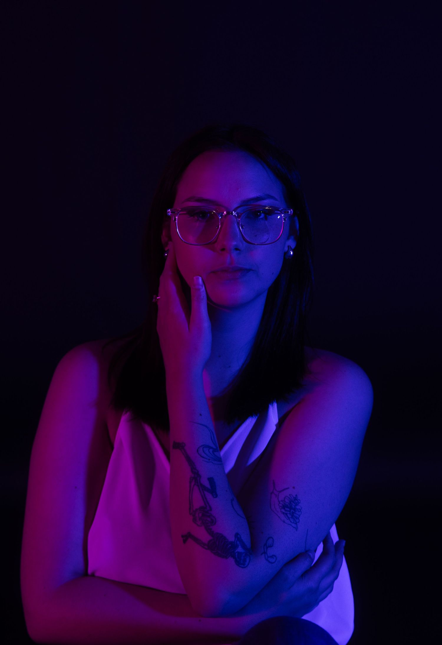 Woman with glasses and tattoo posed thoughtfully under dramatic purple and blue neon studio lighting.