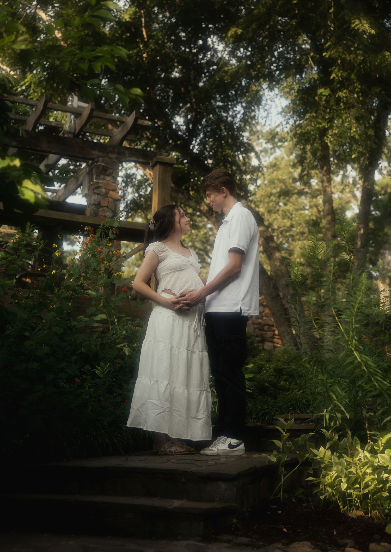 Couple sharing a tender maternity moment on garden steps surrounded by lush greenery and wooden trellis.