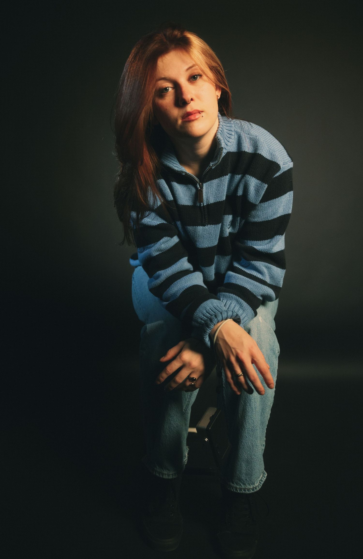 A redheaded woman in a blue striped sweater and jeans poses confidently in a dramatic studio portrait.