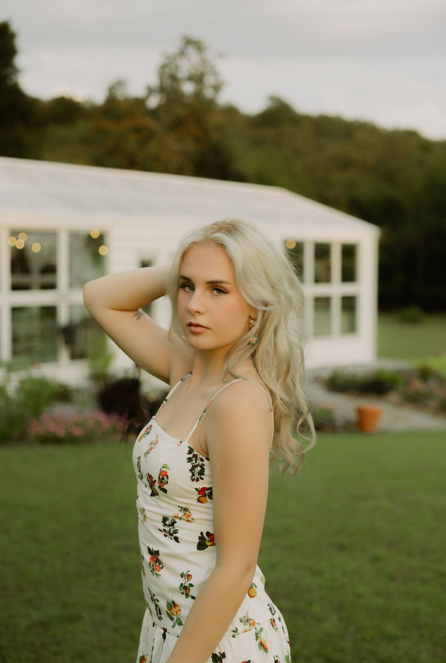 A blonde woman in a floral sundress poses gracefully on a lawn near a white greenhouse.