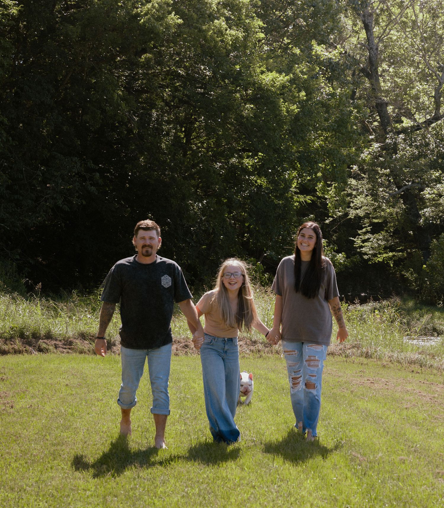 Family of three holding hands walking barefoot across a lush green lawn surrounded by trees.