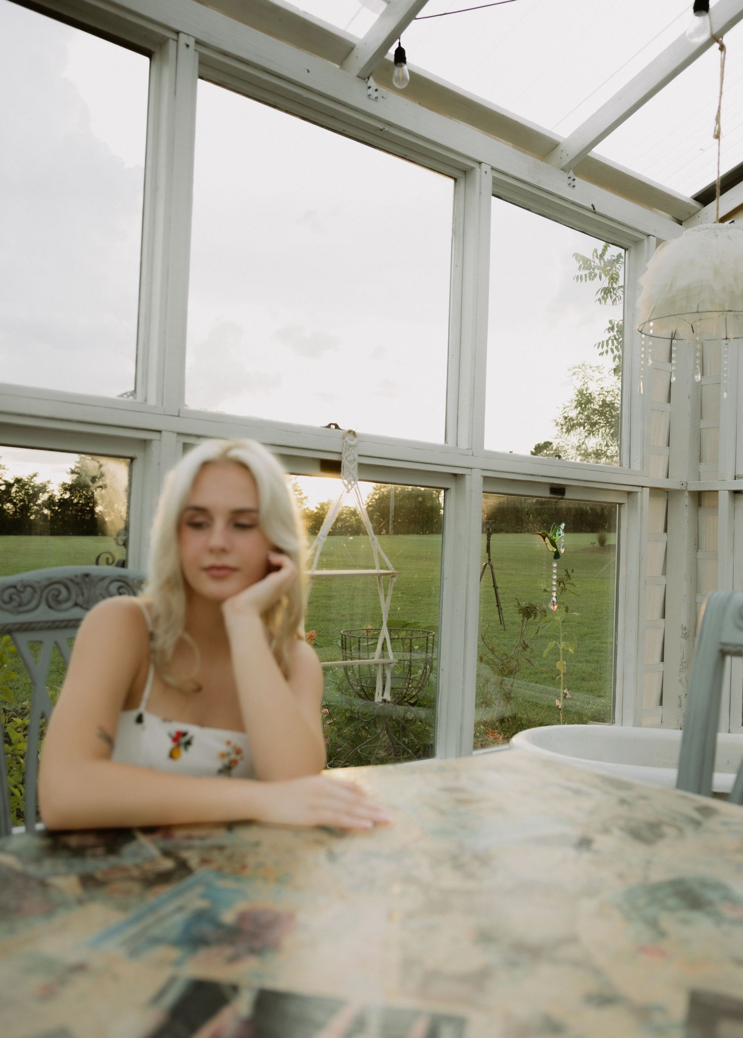 A blonde woman sits pensively at a table inside a bright greenhouse with garden views.