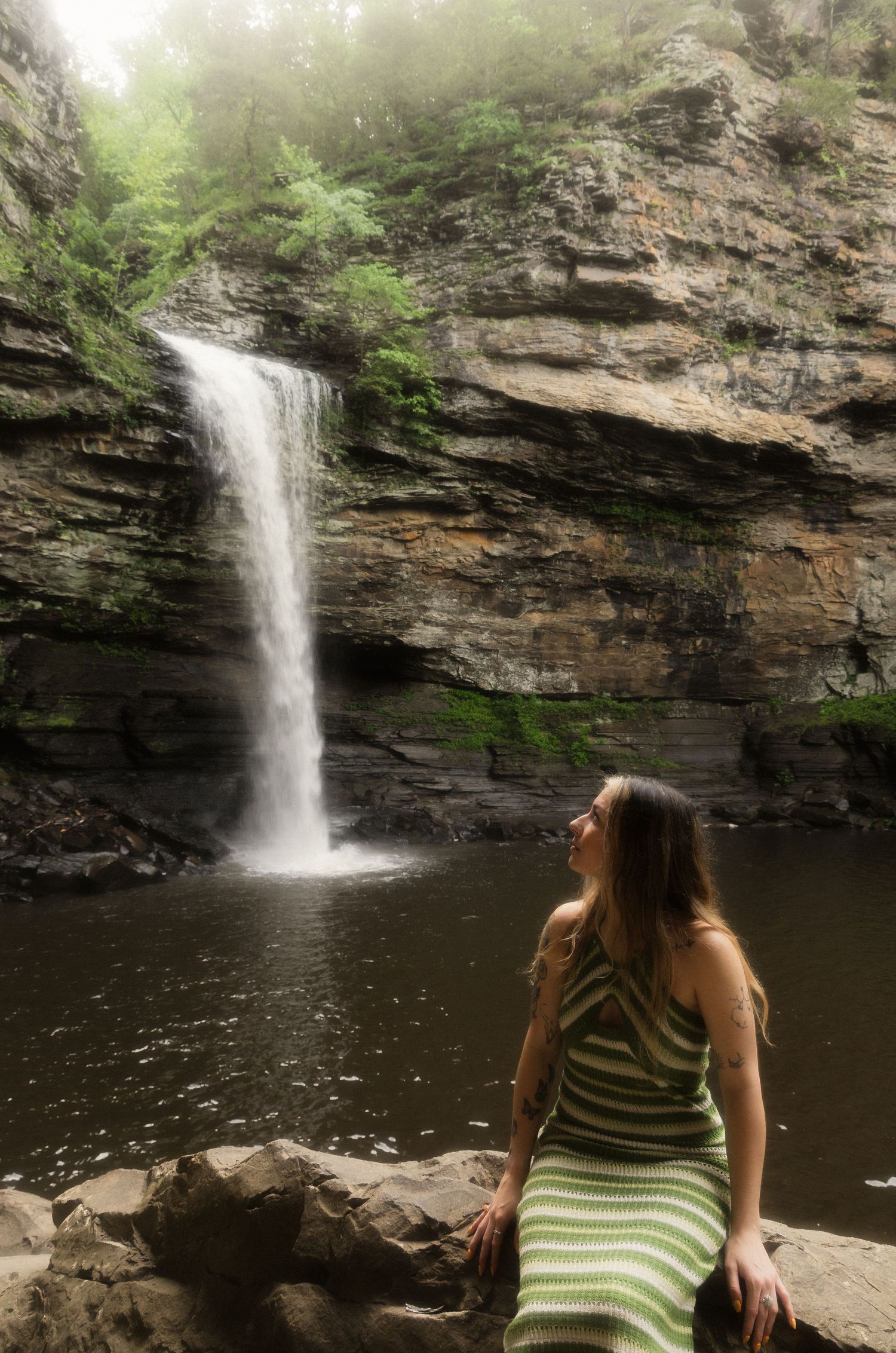 Woman in green striped dress sitting on rocks gazing at misty waterfall in rocky canyon.