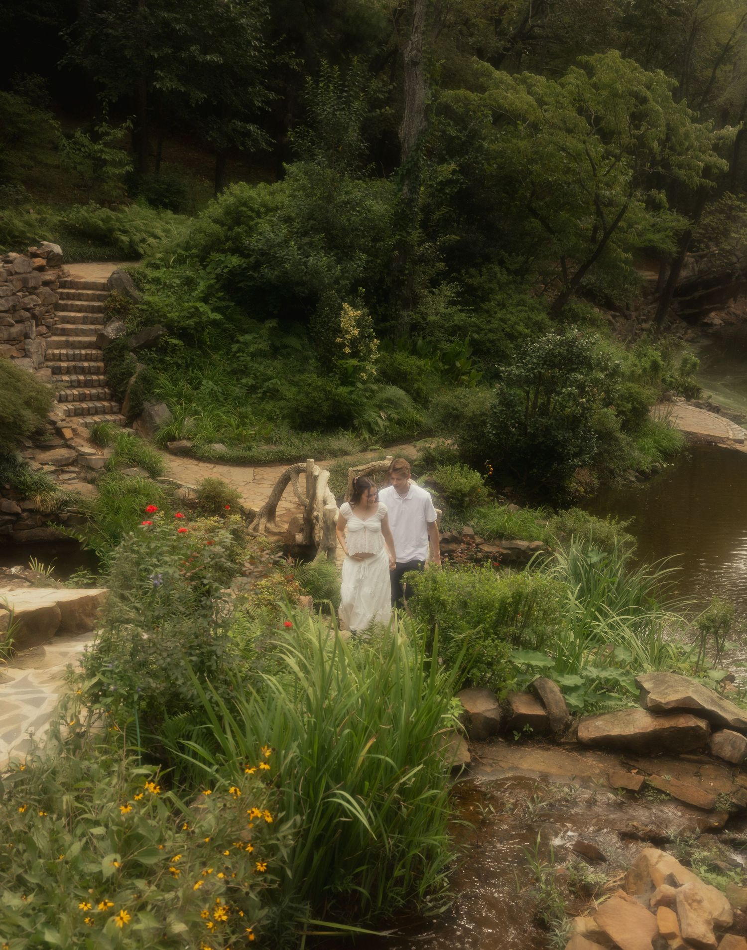 Couple in white attire walks through a lush green garden with stone steps and wildflowers.