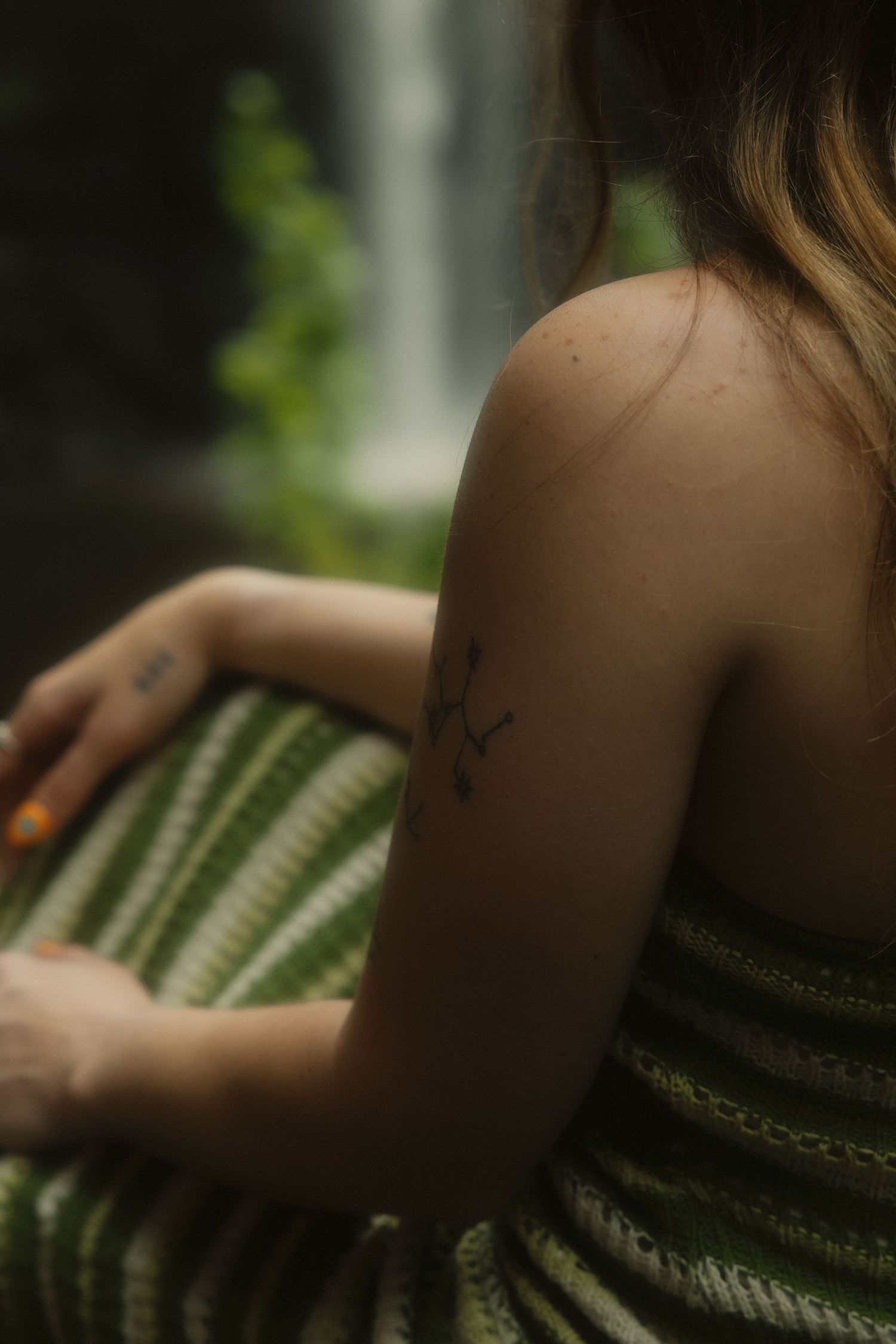 Close-up of woman's shoulder with small branch tattoo, green striped dress, waterfall background.