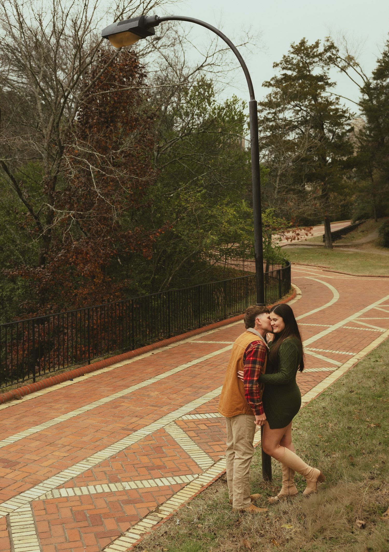 Couple sharing a kiss under a street lamp on a brick pathway in an outdoor park setting.