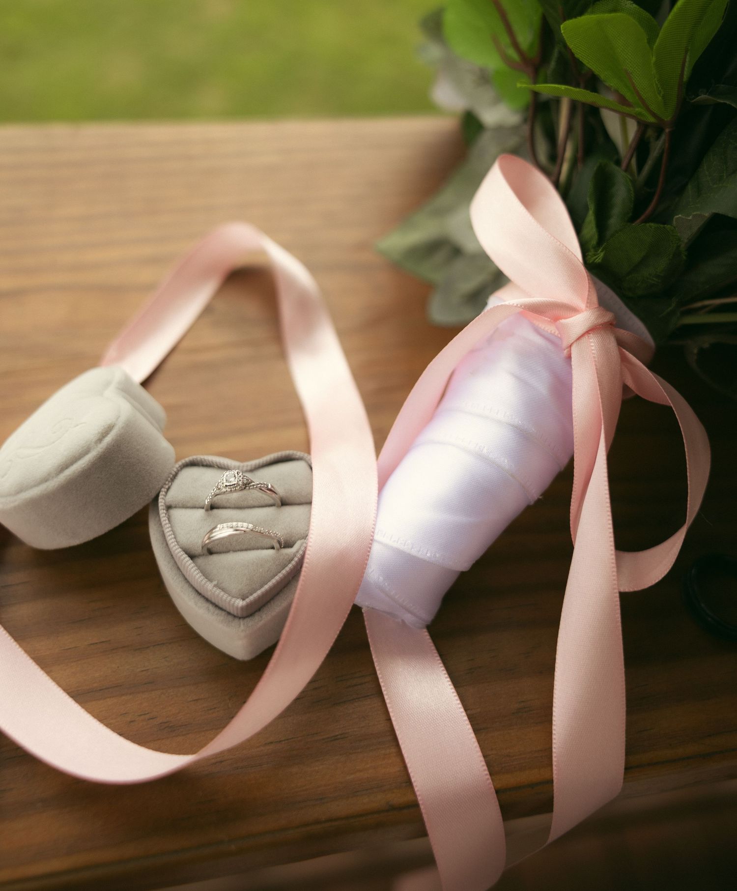 Two wedding rings in a gray heart-shaped velvet box with a flowing blush pink ribbon and green foliage on a rustic wooden table.